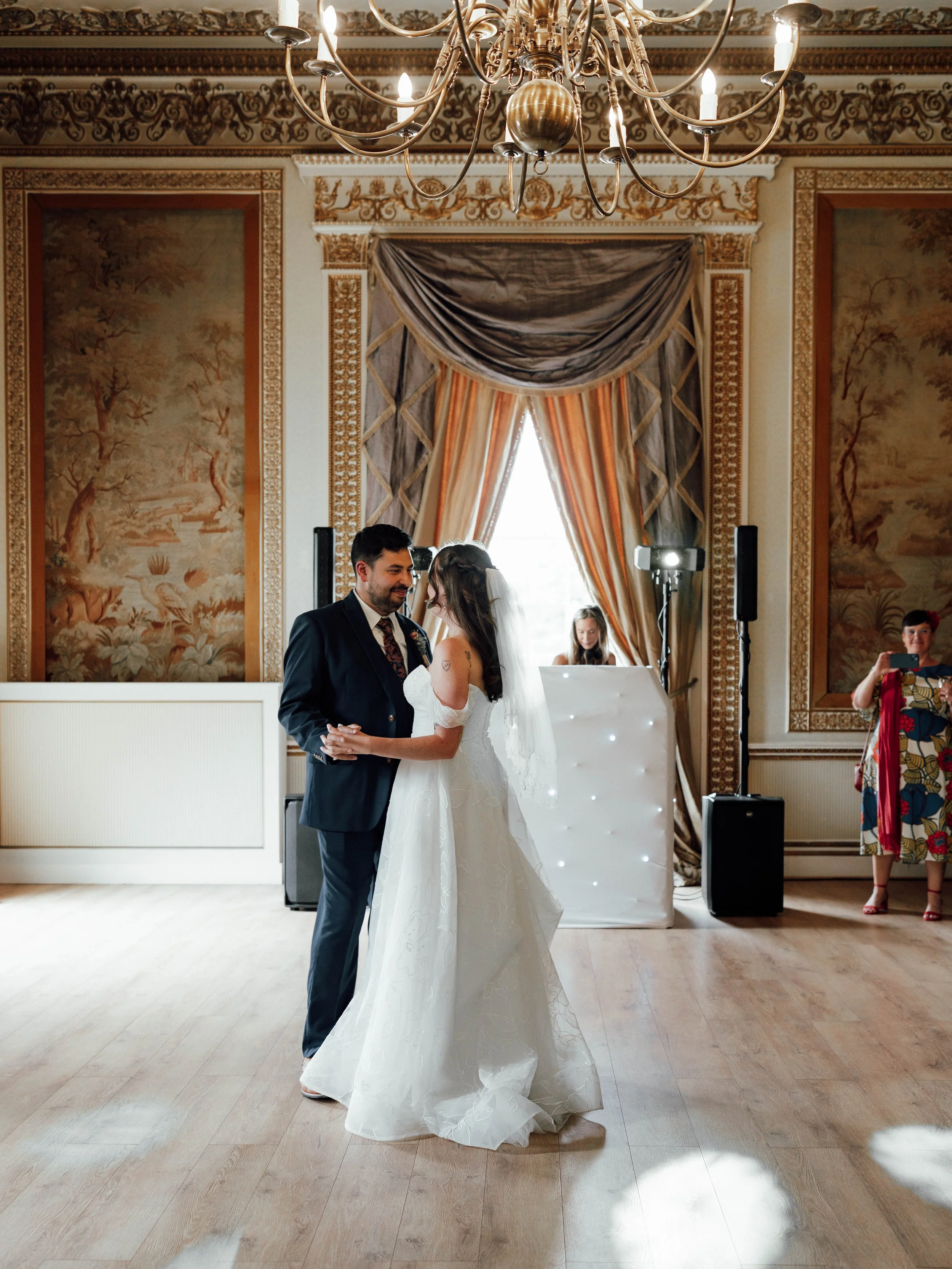 A bride and groom dancing in a decorated wedding hall with a chandelier, ornate wall decor, and large curtains. A woman is taking photos on the right, and a DJ is behind the couple.