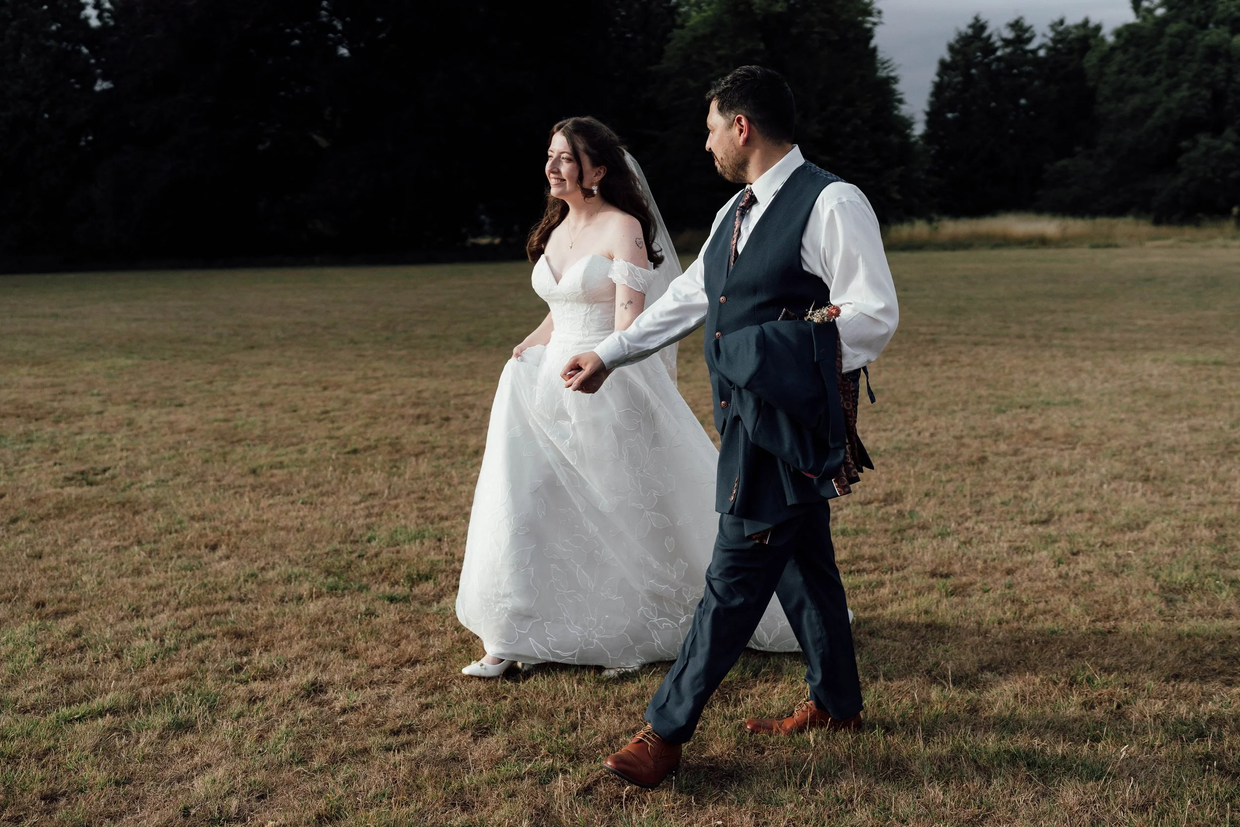 A bride wearing a white wedding gown walking on a grassy field, holding her dress with a smiling man in a navy blue vest and white shirt, walking beside her on a cloudy day.