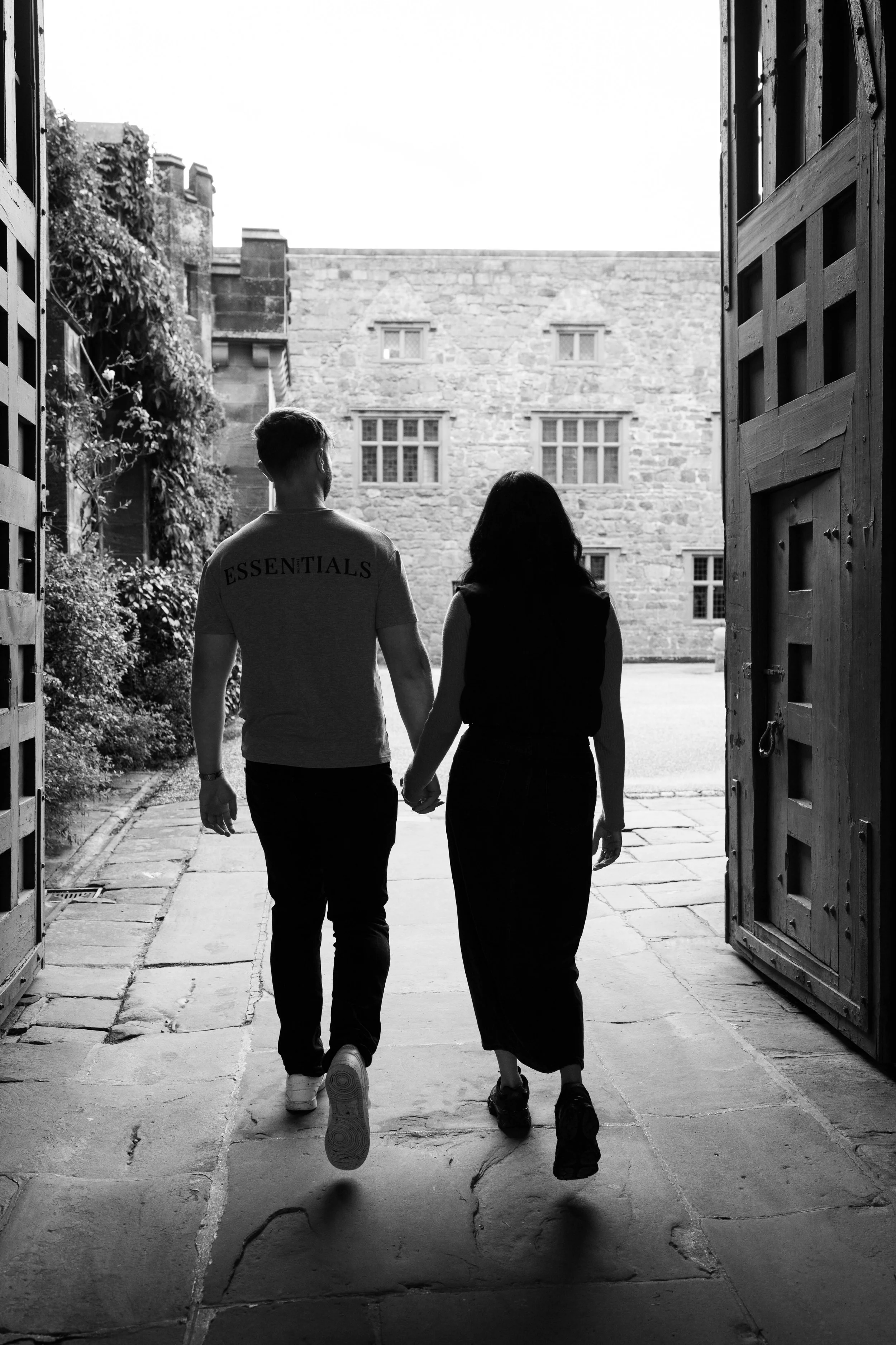 A black and white photo of a young man and woman holding hands as they walk out of a wooden gate into a courtyard with stone paving and a historic stone building in the background.