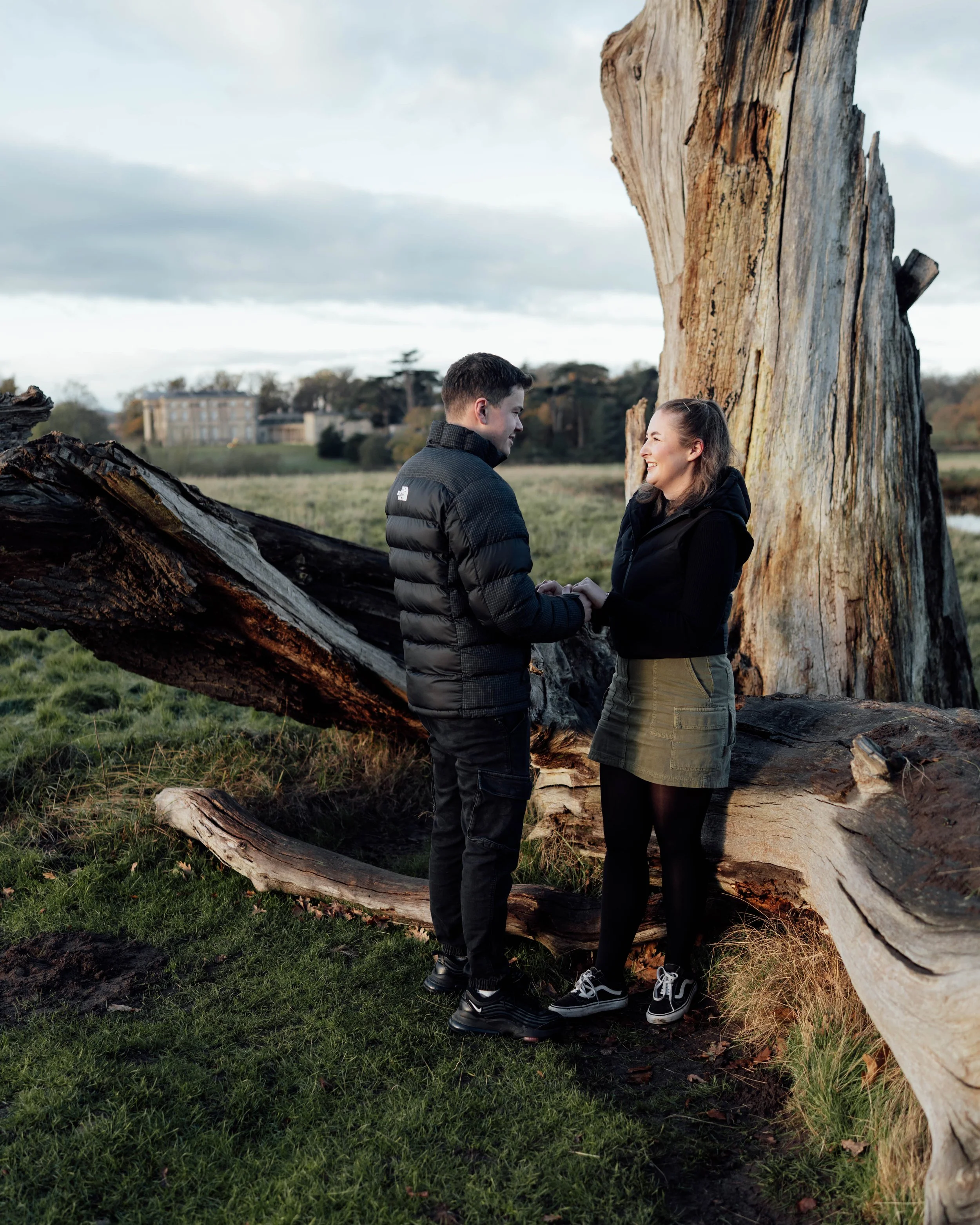 A couple holding hands and smiling while standing outdoors near a large fallen tree and a tall broken tree trunk.