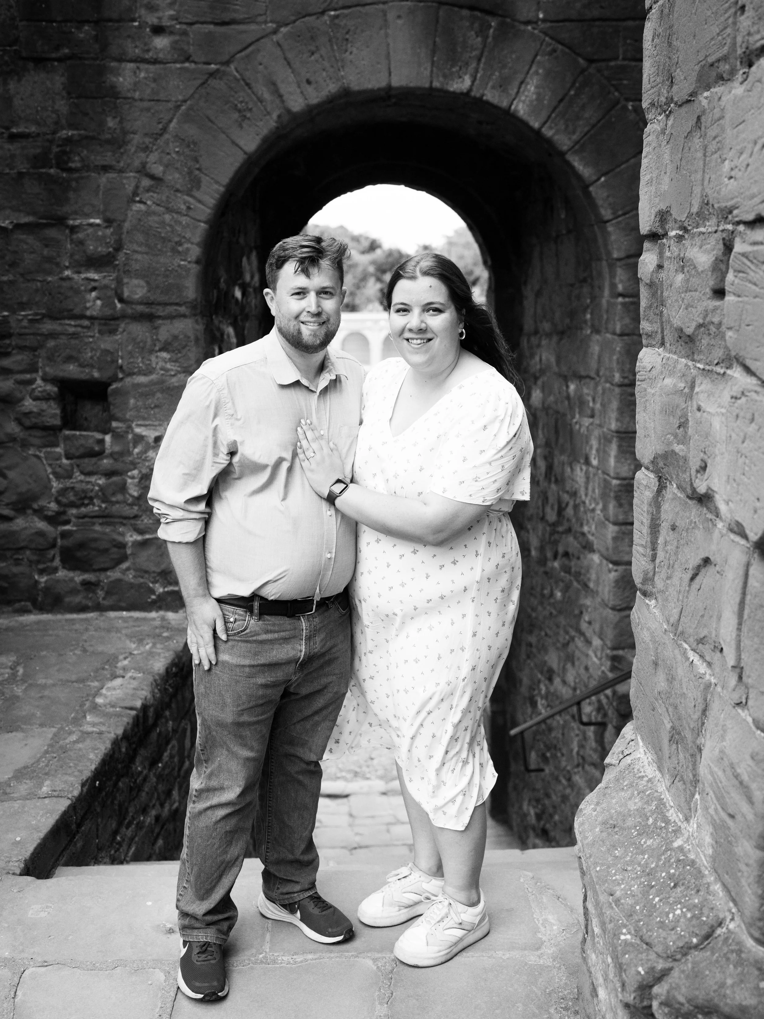 A smiling couple standing on stone steps under an archway with a stone wall on one side, behind them is an outdoor scene.
