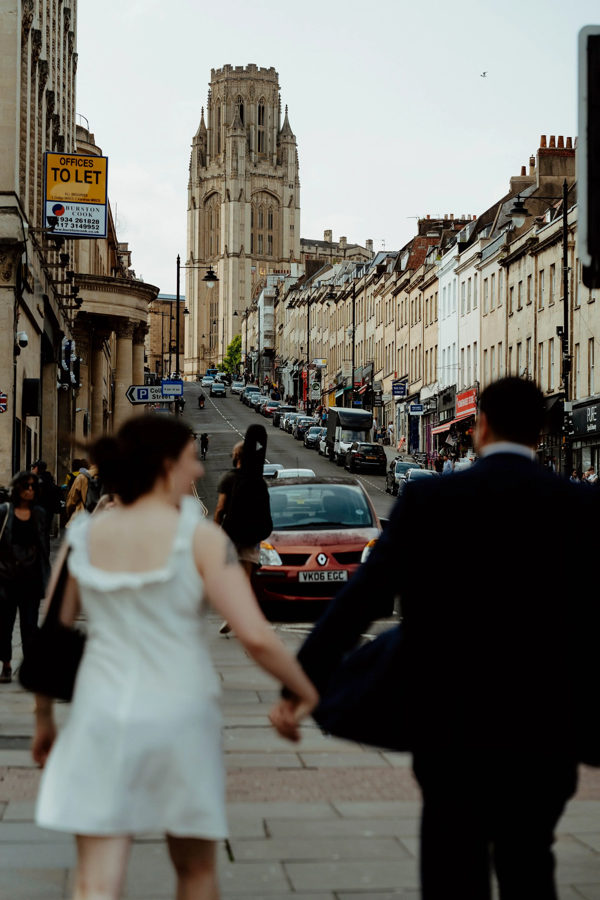 A city street scene with a Gothic church tower in the background, cars parked along the street, and people walking, including a woman in a white dress and a man in a suit holding hands in the foreground.