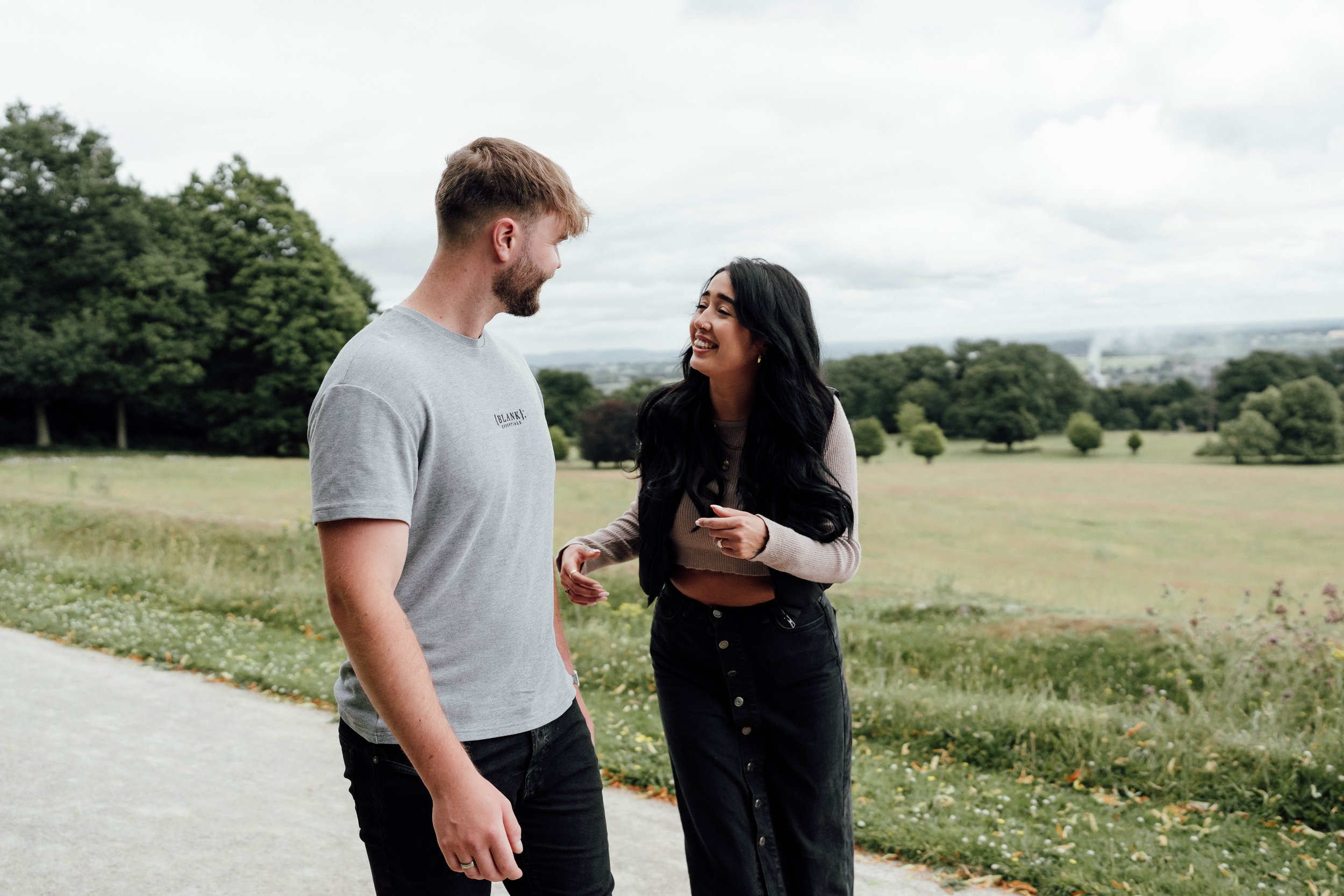A man and woman standing outdoors on a cloudy day, smiling and looking at each other in a park with trees and open fields in the background.