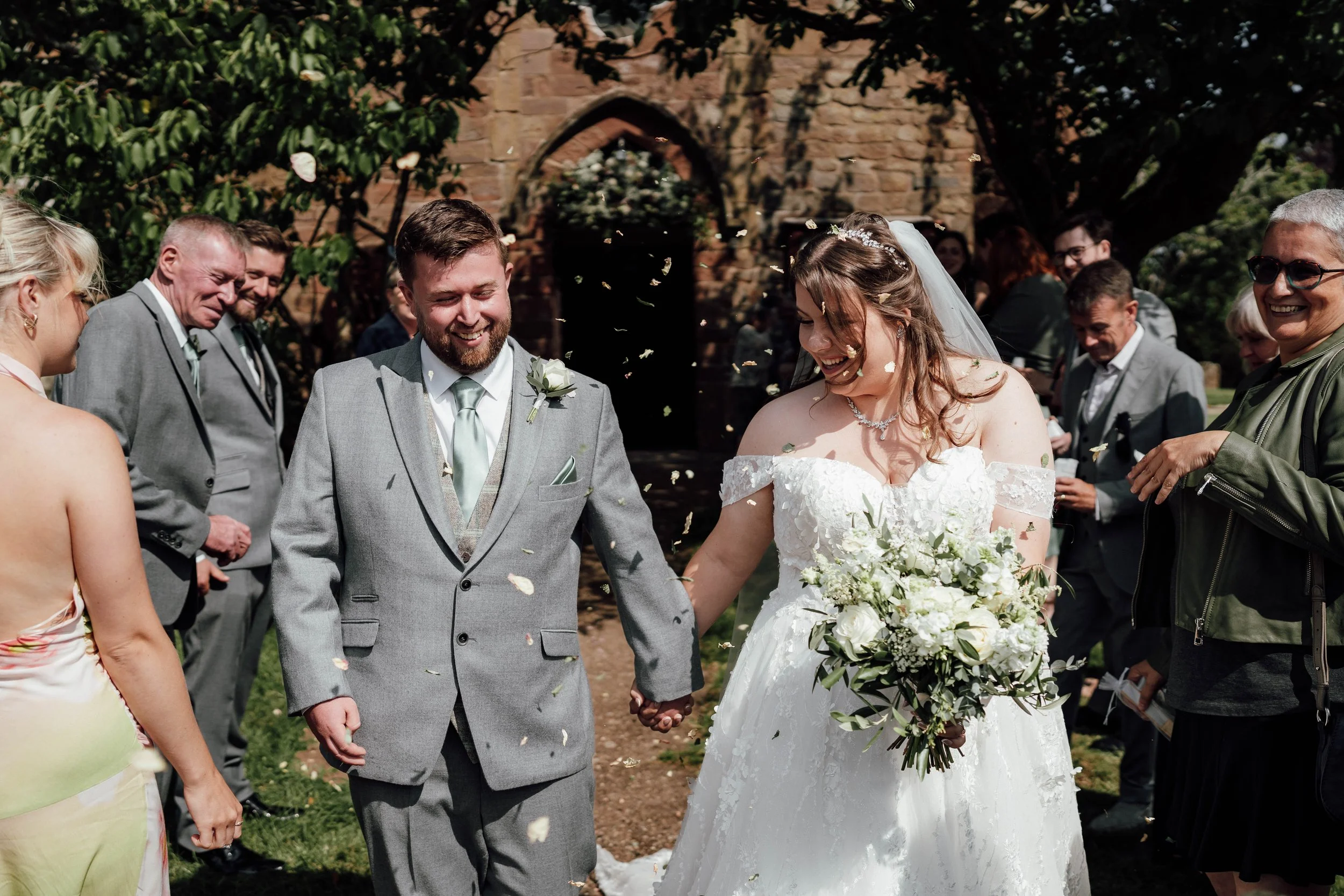 A happy bride and groom celebrating their wedding outside, surrounded by smiling guests, with the bride holding a large bouquet and the groom holding her hand, as confetti falls around them.