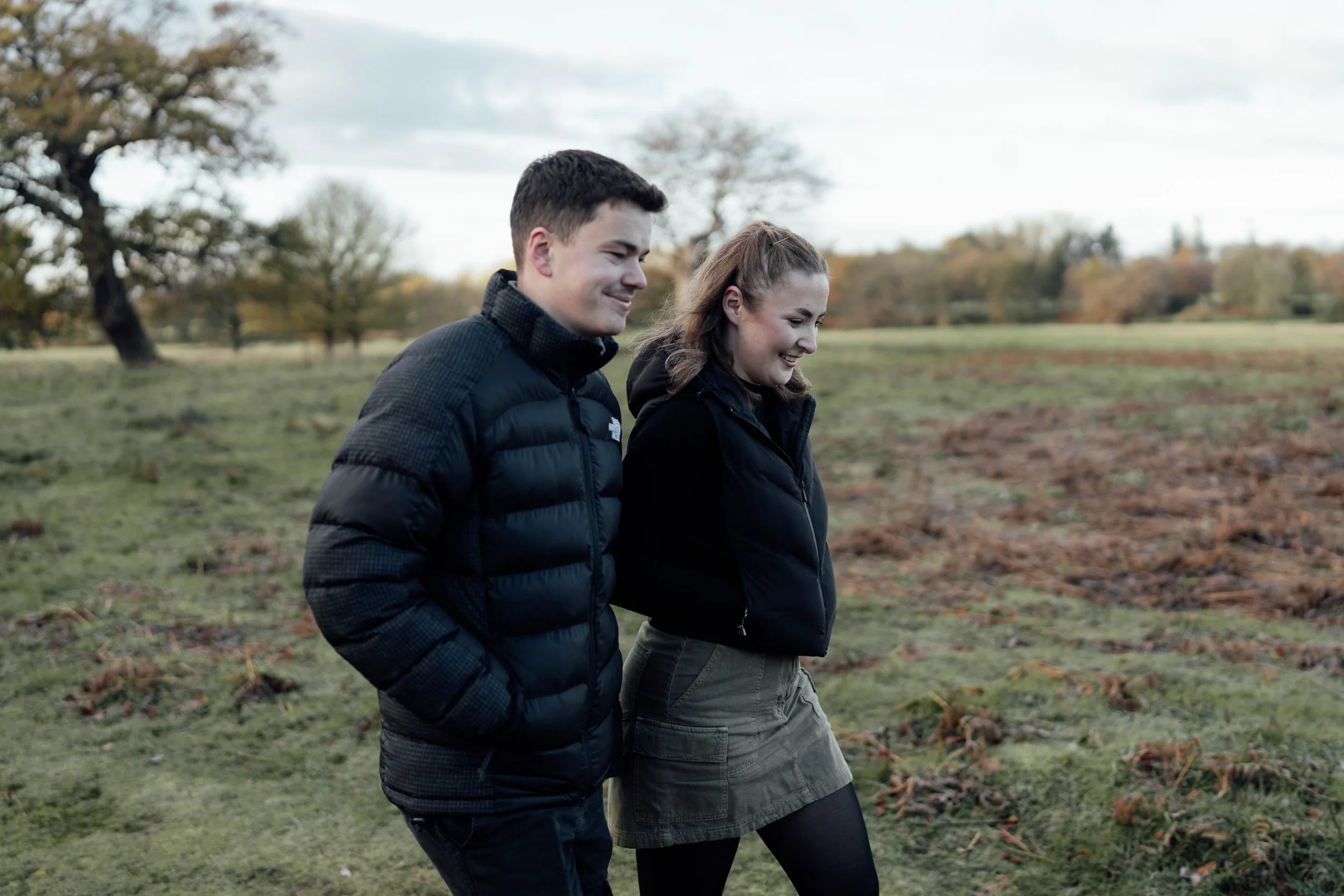 A young man and woman walking in a grassy field during fall, smiling and enjoying each other's company.