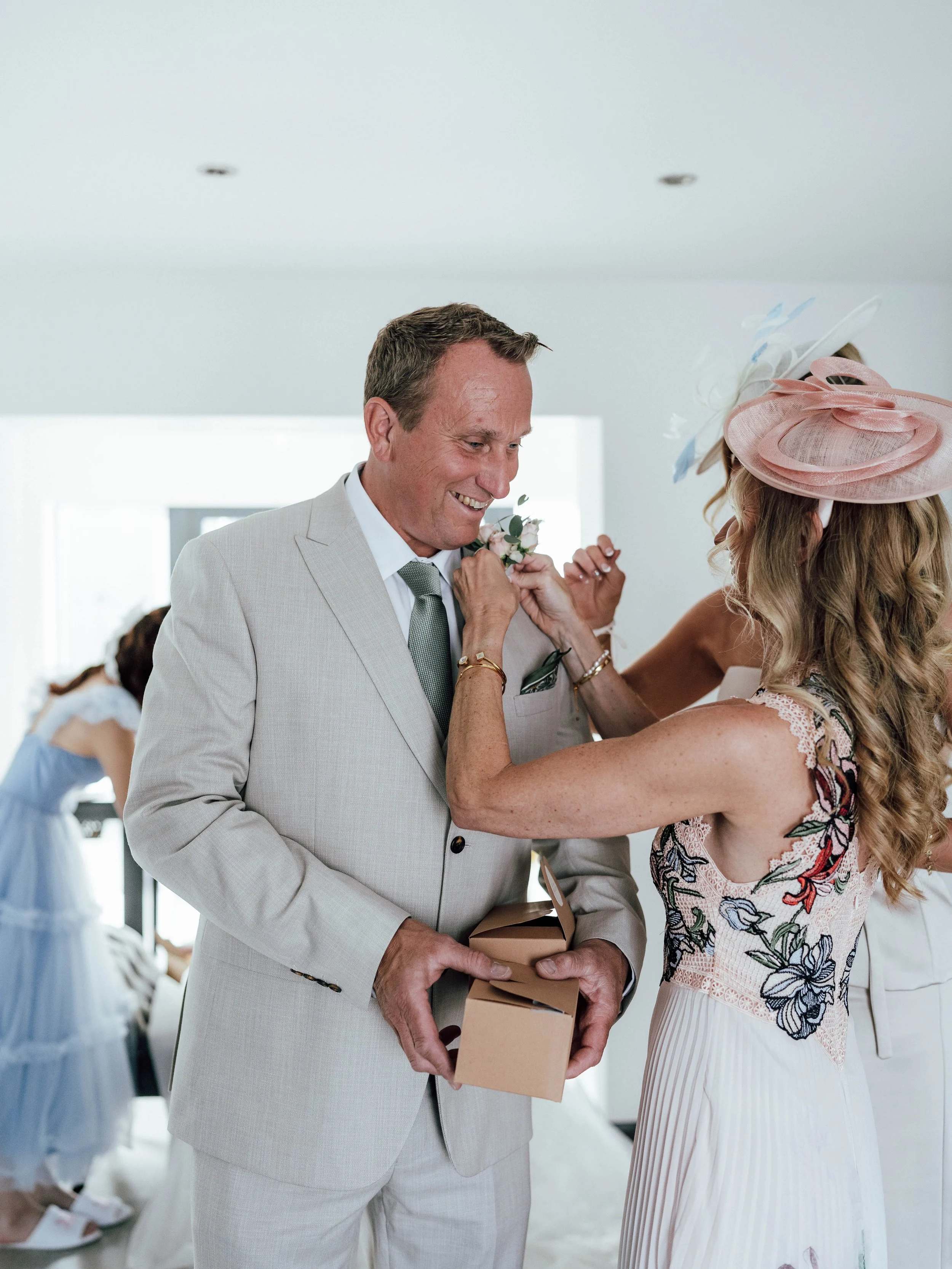 A woman with long, wavy hair and a pink hat pins a boutonniere on a man in a light-colored suit, who is holding a box, during a wedding celebration.