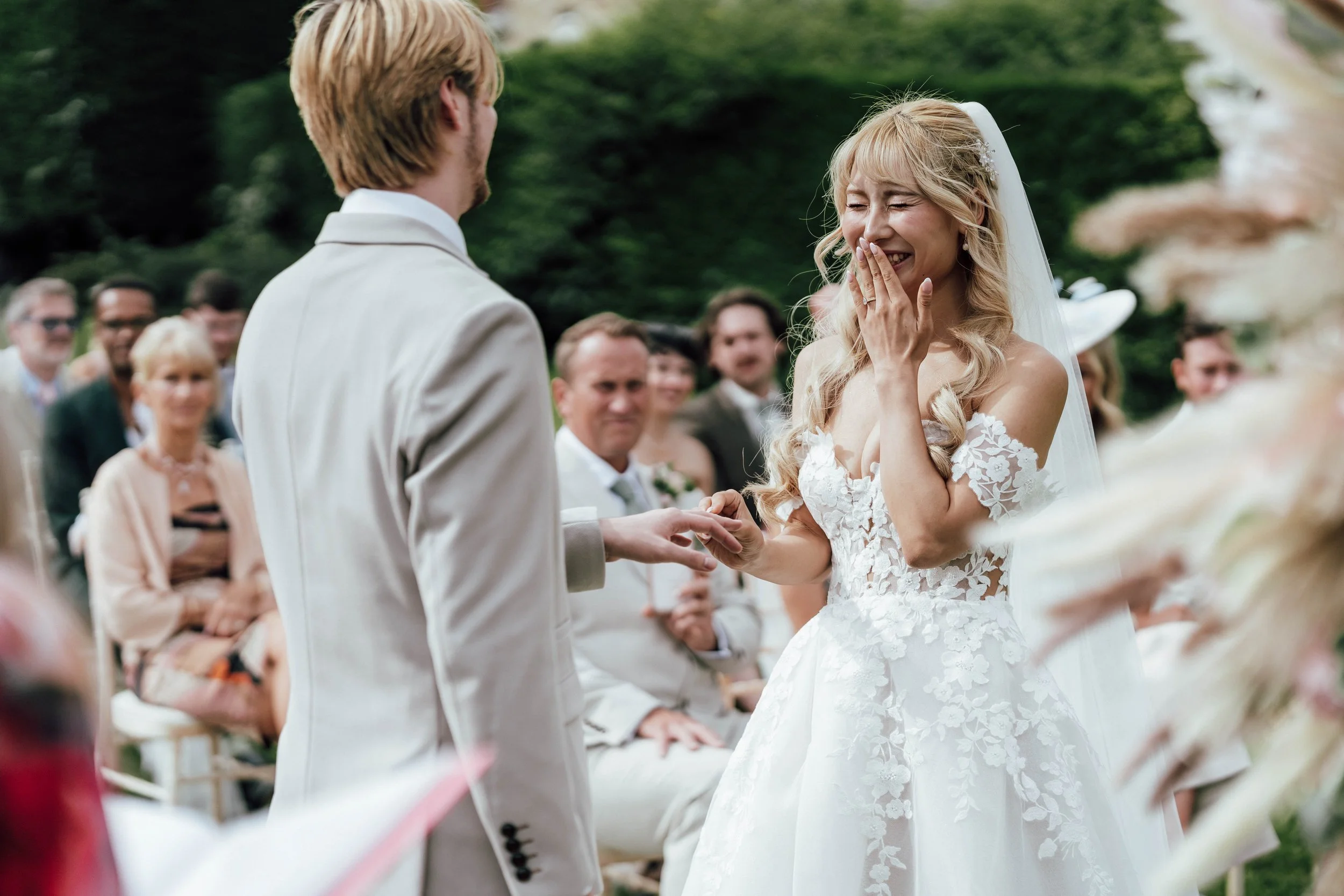 A bride and groom exchanging vows at an outdoor wedding ceremony, with guests seated in the background, surrounded by greenery.