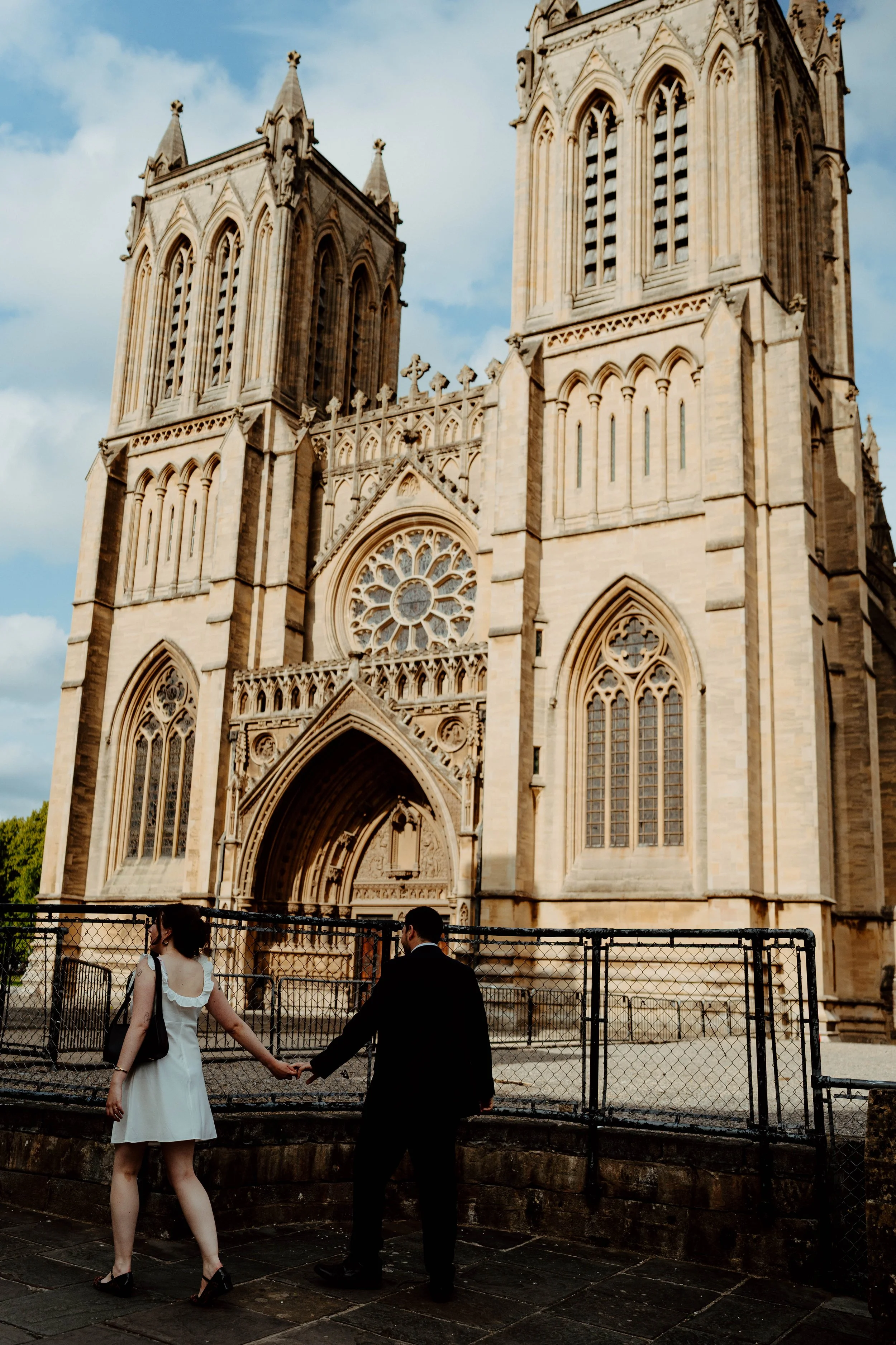 A large, historic Gothic cathedral with two tall towers and detailed stone architecture, featuring arches and a rose window, with a man and woman in formal attire holding hands in the foreground.
