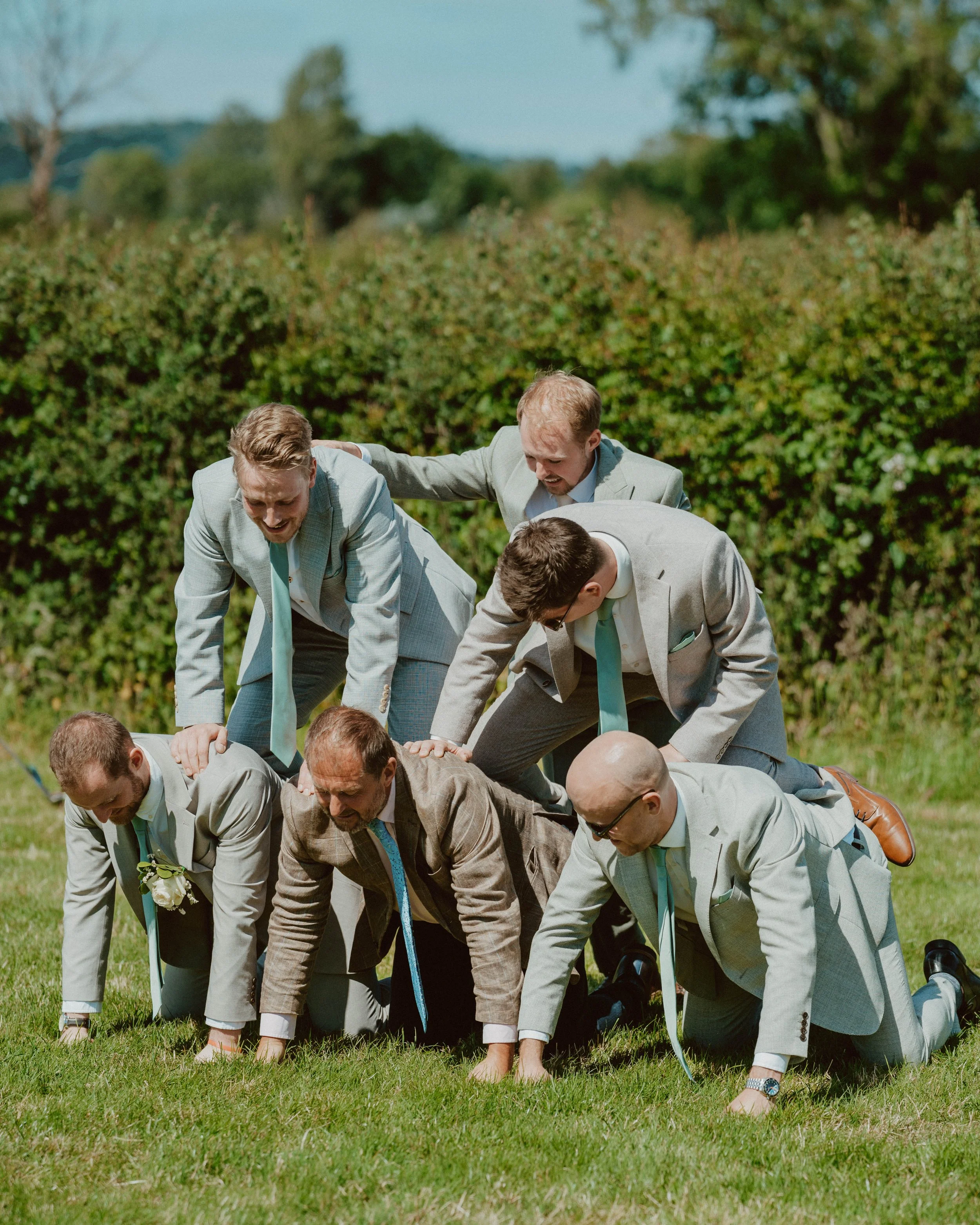 Group of men dressed in suits stacking on top of each other outdoors on grass, forming a human pyramid, with trees and bushes in the background.