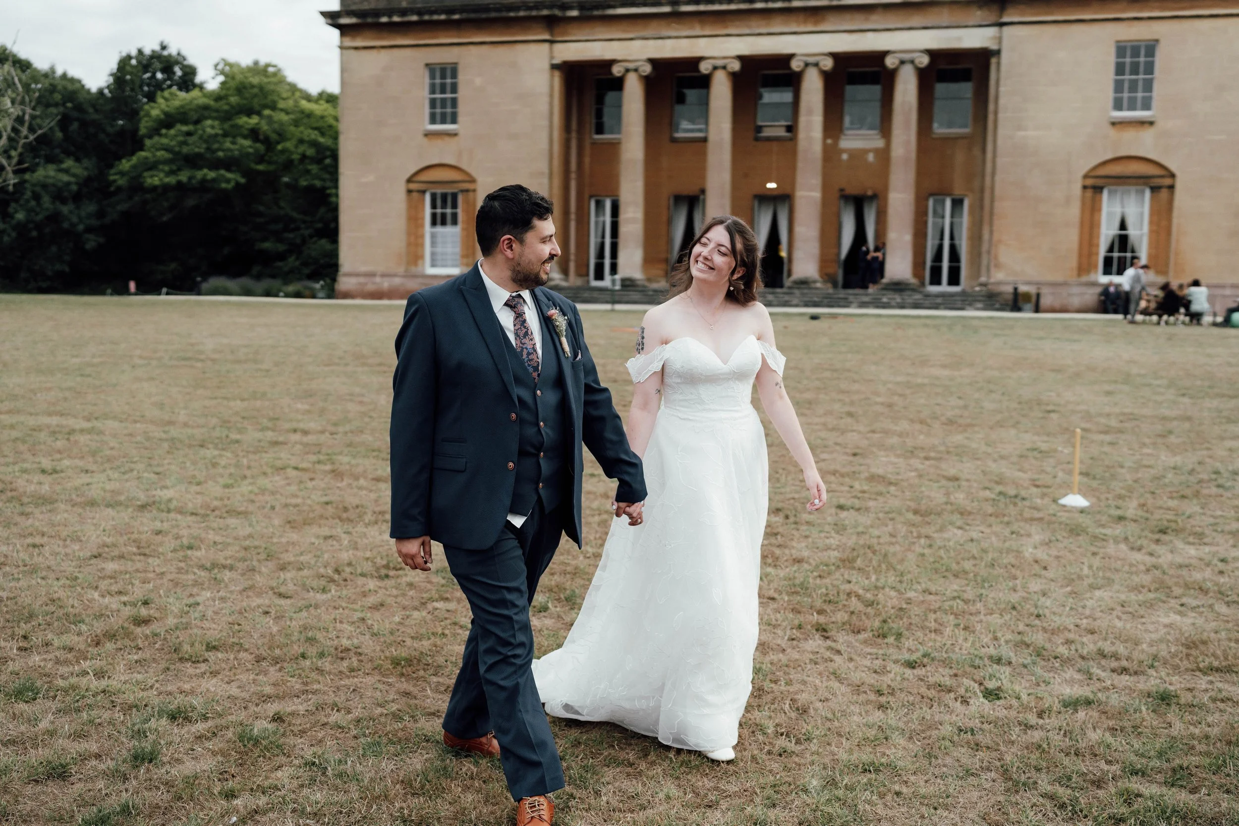 Bride and groom holding hands, walking on a grassy lawn in front of a large historic building, smiling at each other.