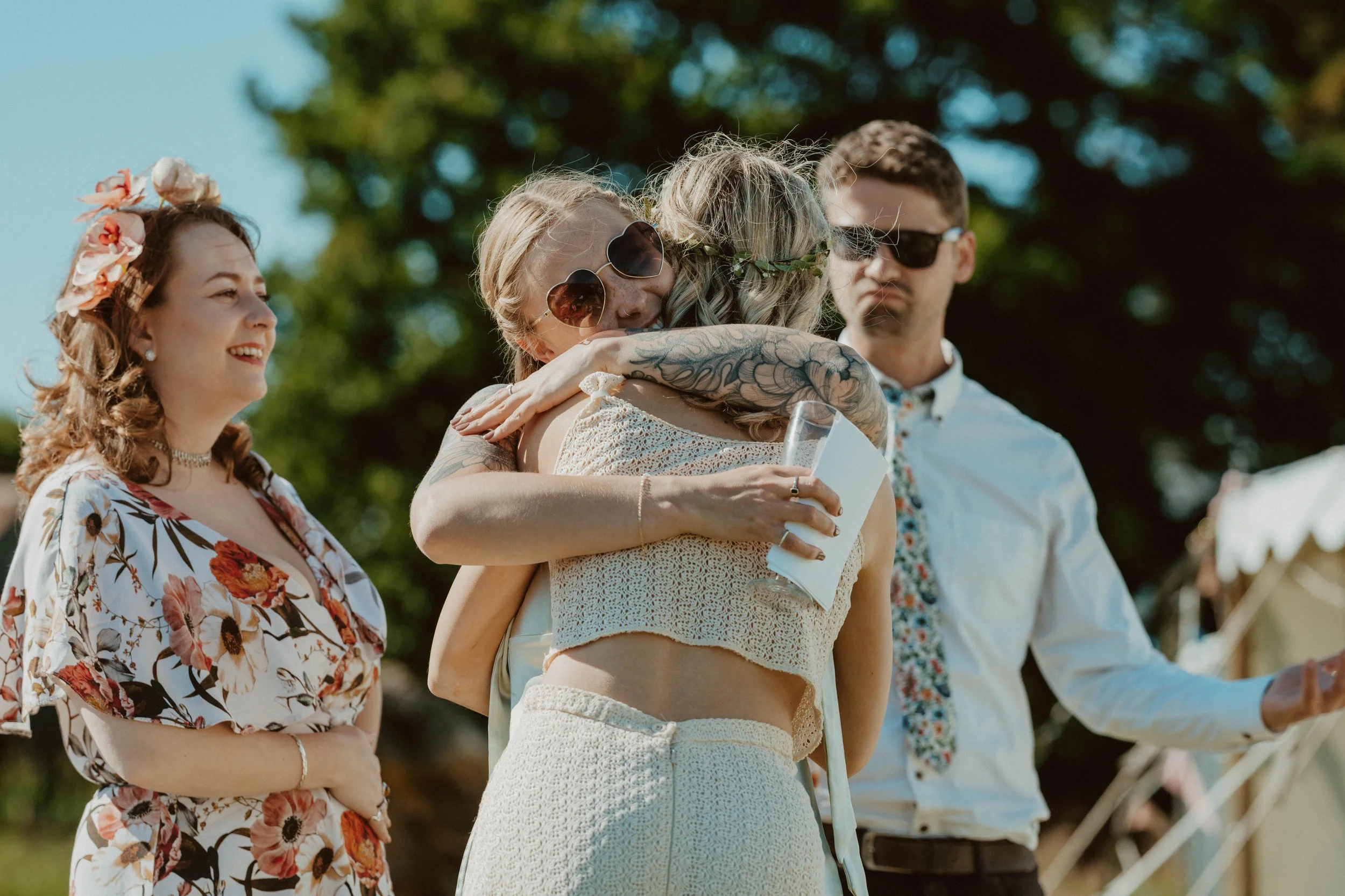 People hugging outdoors during a sunny event, wearing summer clothes and sunglasses, with tents and trees in the background.