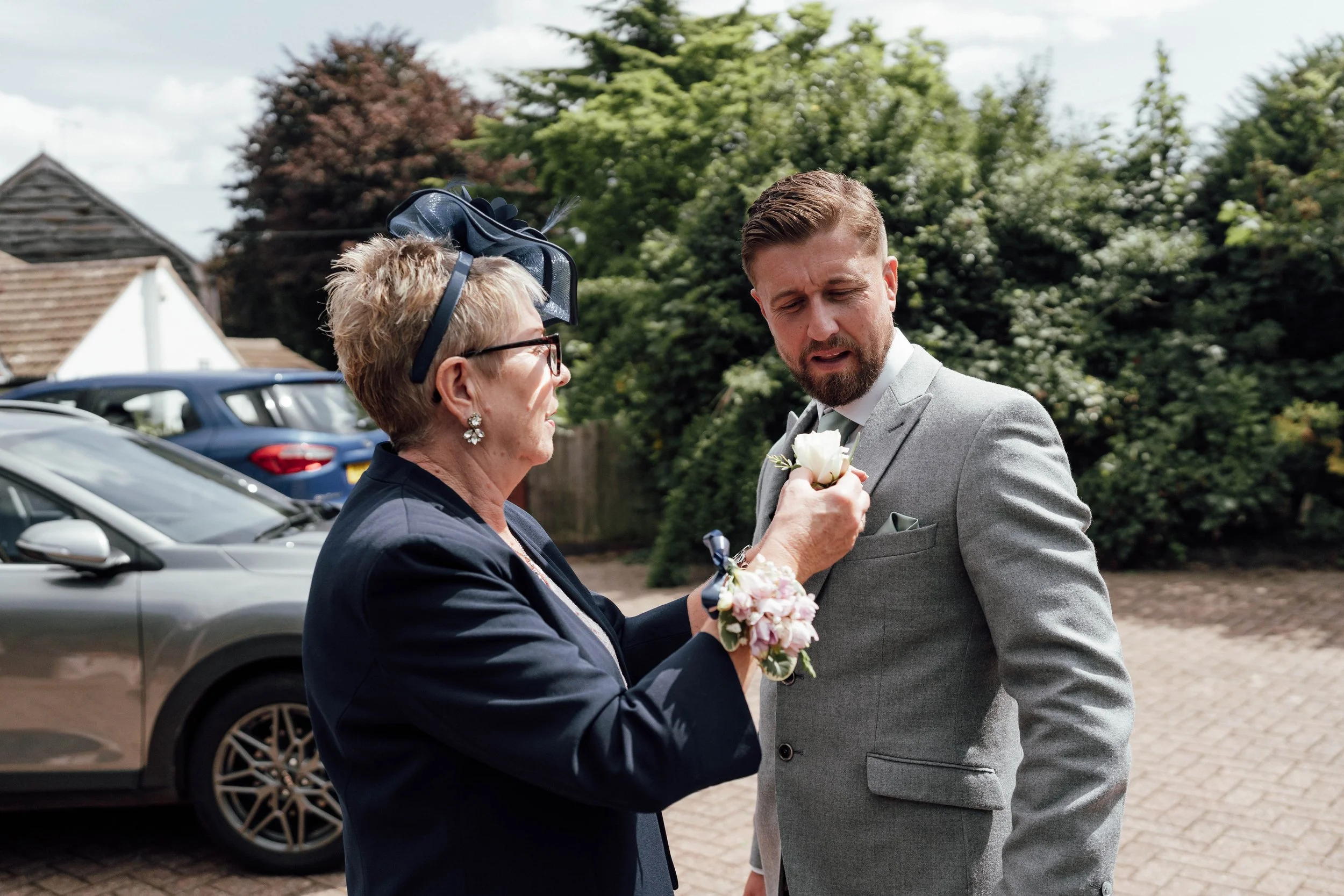 An elderly woman and a young man are outdoors near parked cars, with trees and houses in the background. The woman is pinning a white flower onto the man's gray suit. The woman is wearing glasses, a dark blazer, and a decorative headpiece, while the 