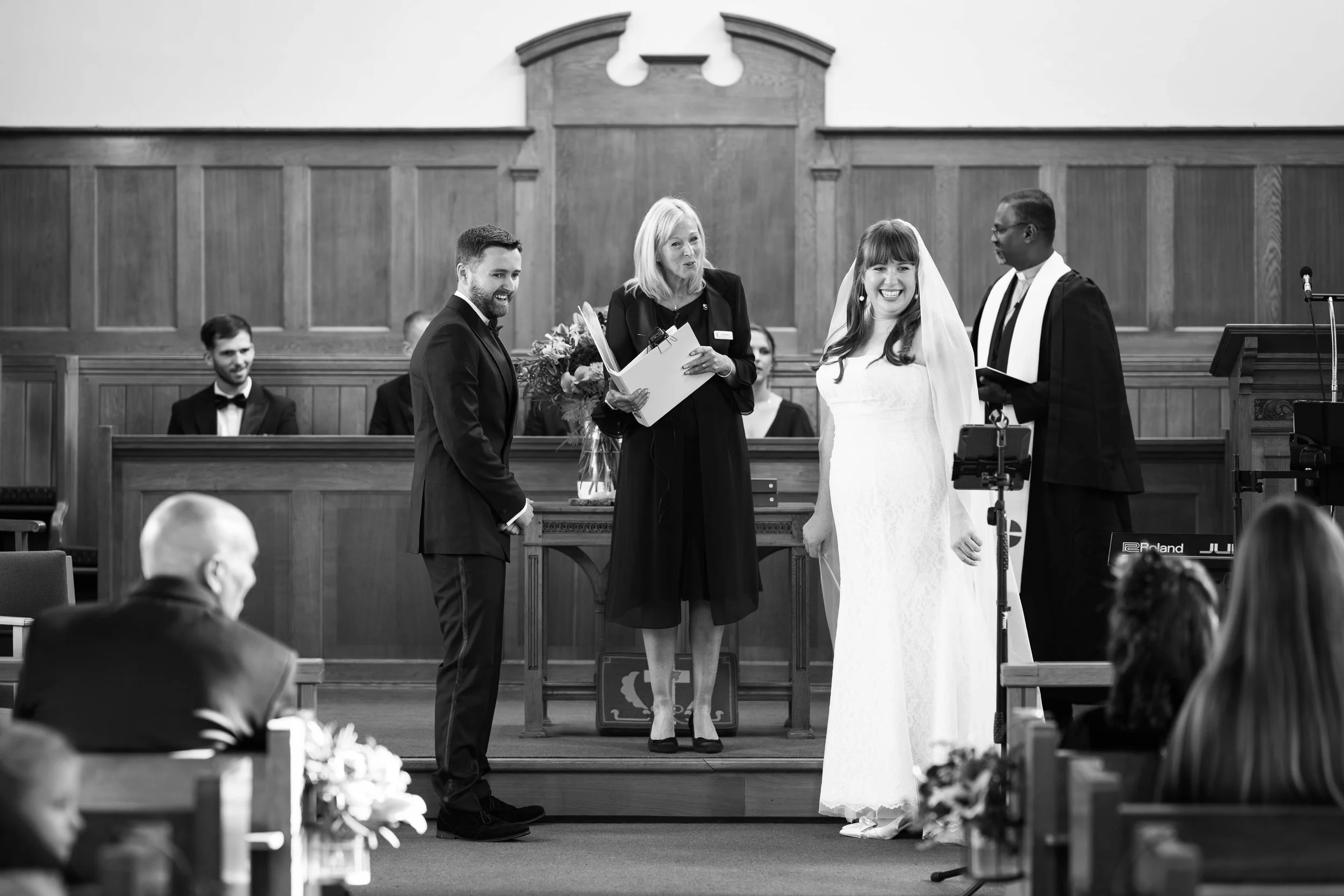 A wedding ceremony inside a church with a bride in a white dress and veil, groom in a suit, officiant holding papers, and a pastor in robes standing at an altar with other people seated behind.