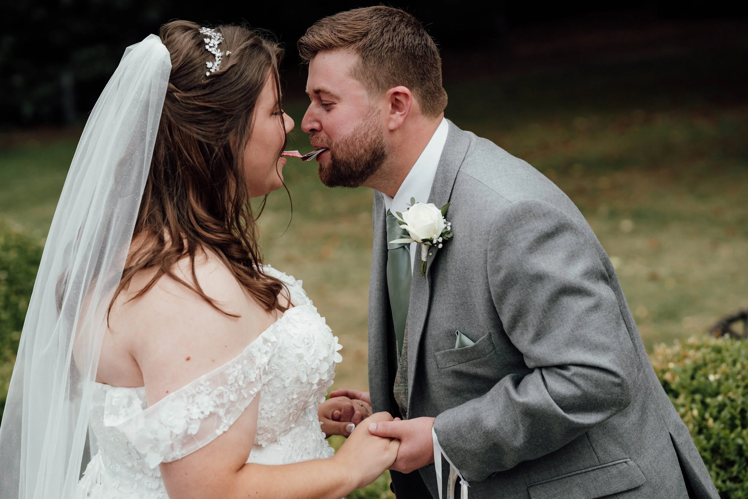A bride and groom with their foreheads touching, holding hands, and lollipops in their mouths during a wedding ceremony outdoors.