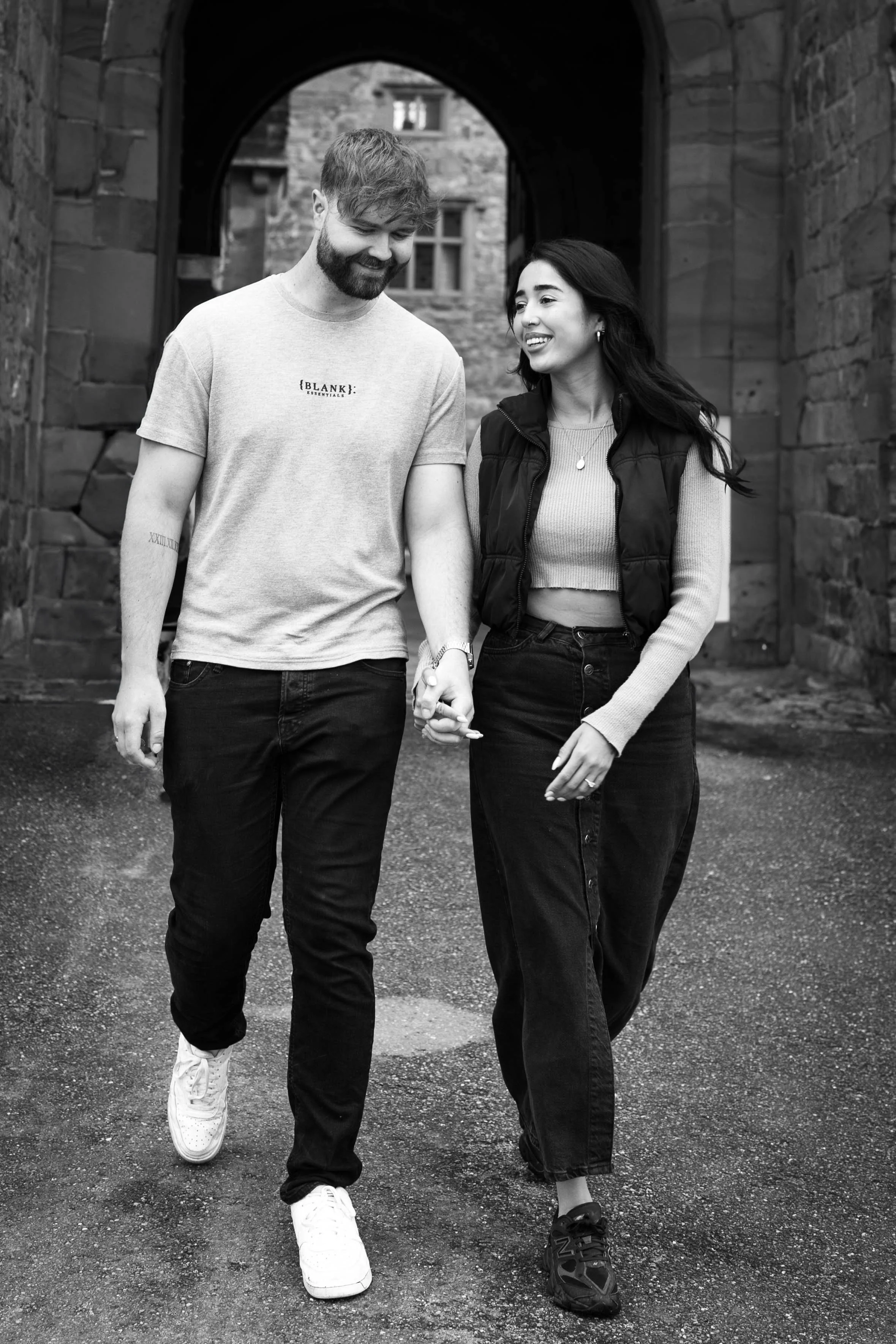A couple walking hand-in-hand outdoors, smiling and looking at each other, on a paved path with stone building walls and an archway in the background.