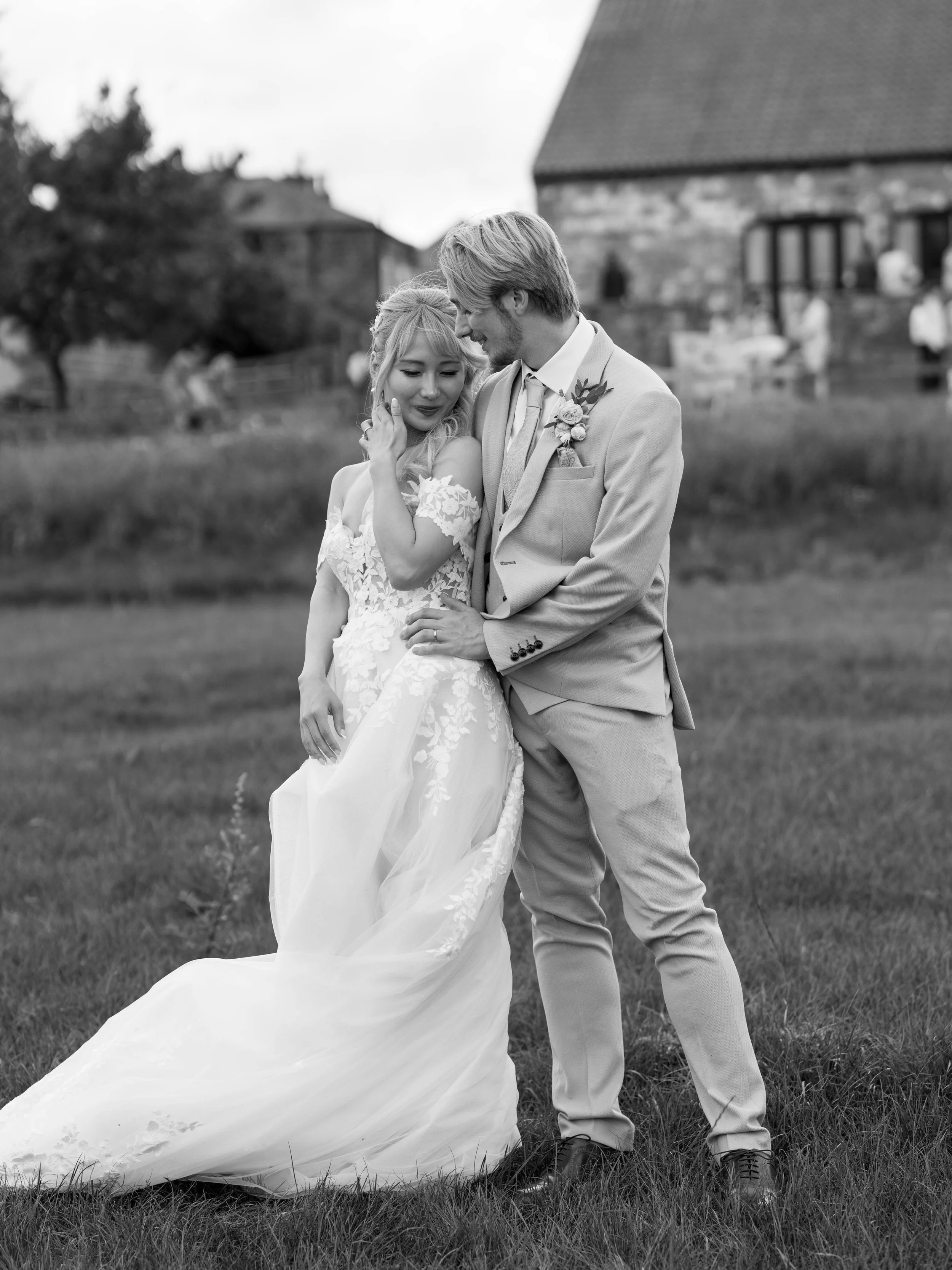A black and white photo of a bride and groom standing on grass, embracing each other, outdoors with a rustic building and people in the background.