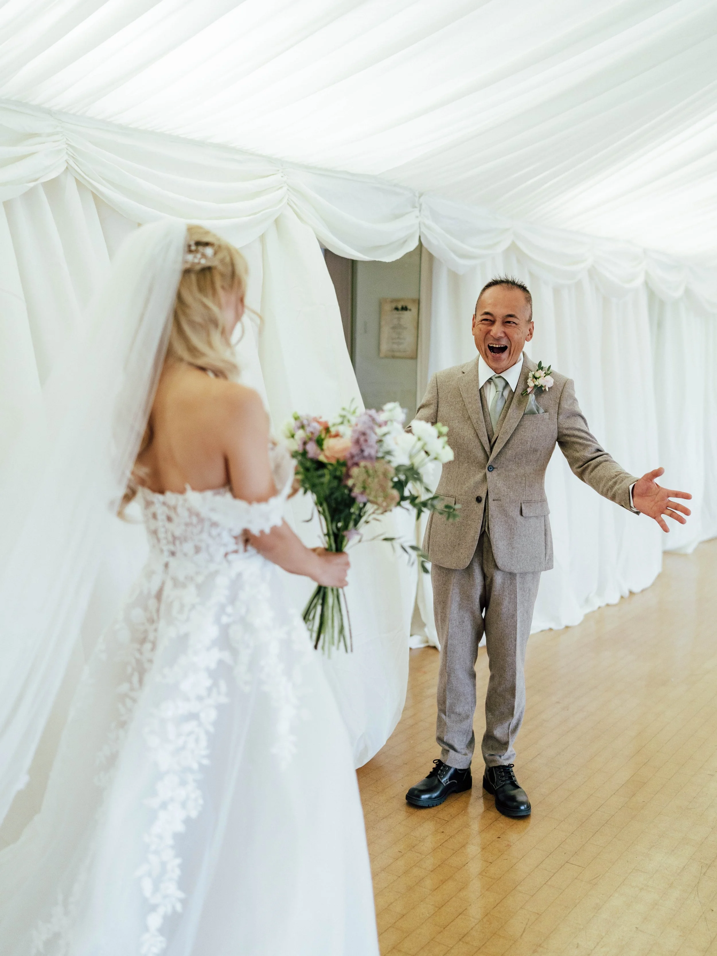 A bride holding a bouquet of flowers stands facing a man in a grey suit who is smiling and gesturing at her inside a decorated white tent.