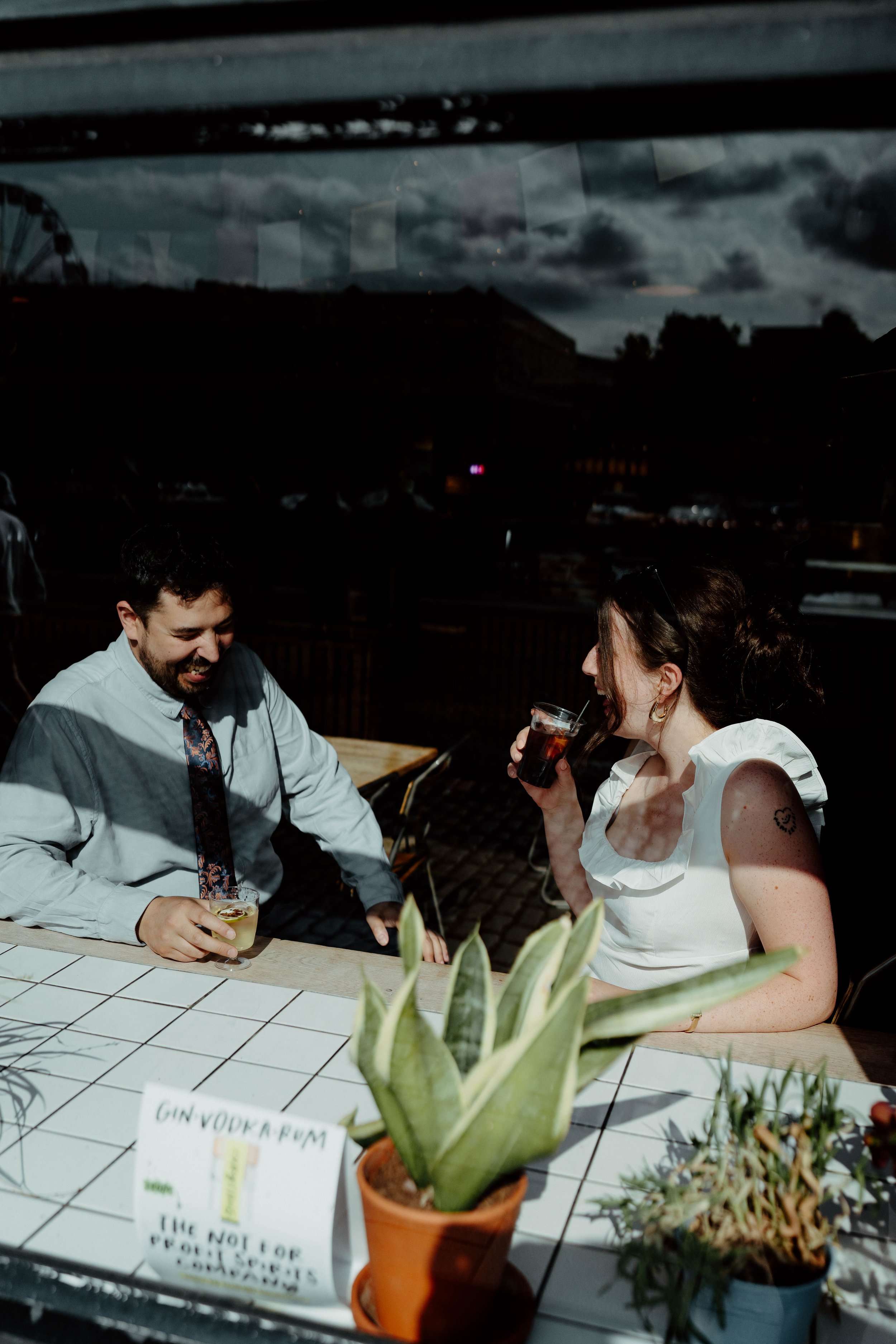 Two people, a man and a woman, sitting at an outdoor table enjoying drinks at night with a dark and cloudy sky in the background.