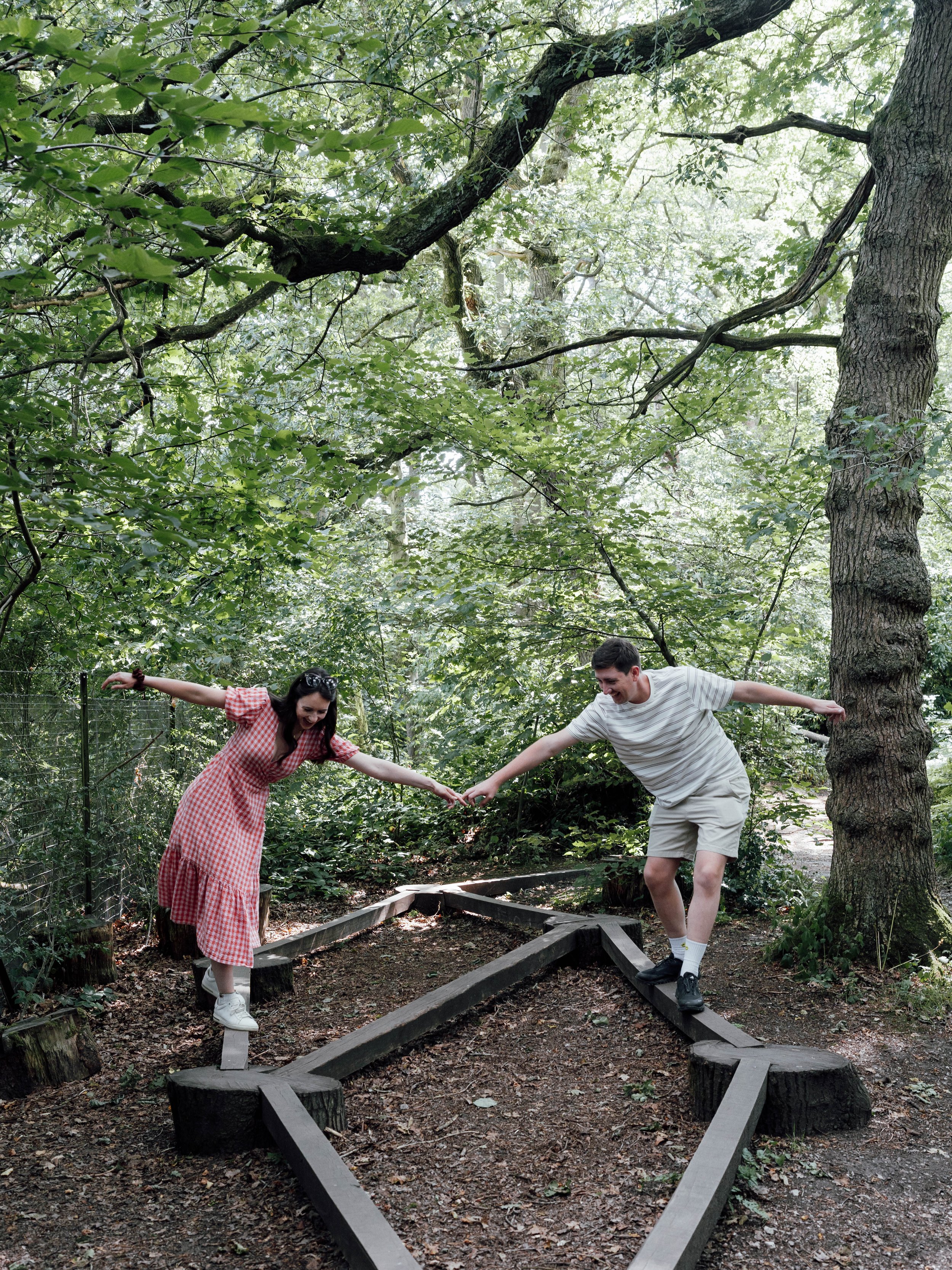 A young man and woman are balancing on wooden planks in a forest, holding hands and smiling.