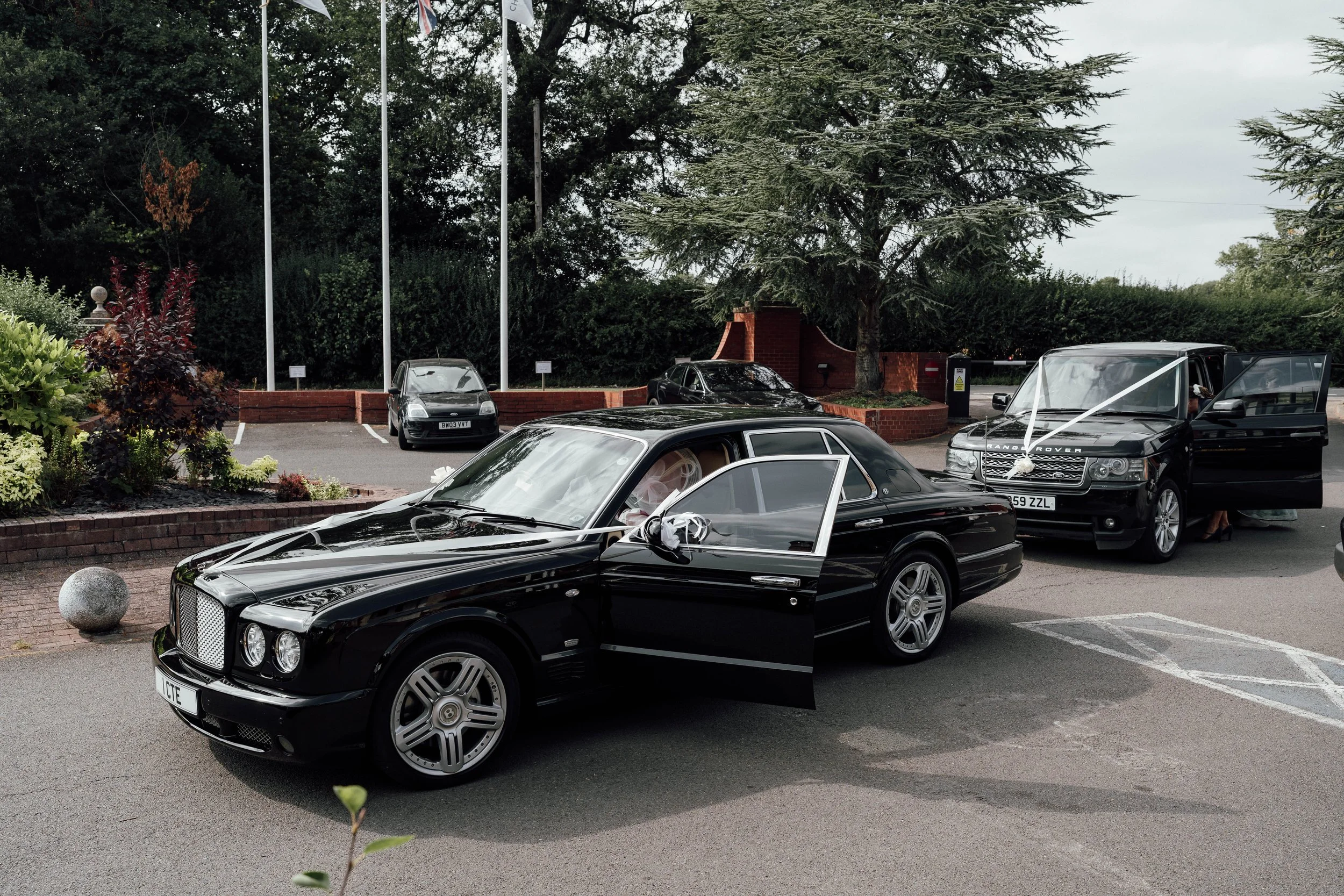 Black luxury car with wedding ribbons parked outside during daytime, with a Range Rover and other vehicles in the background.