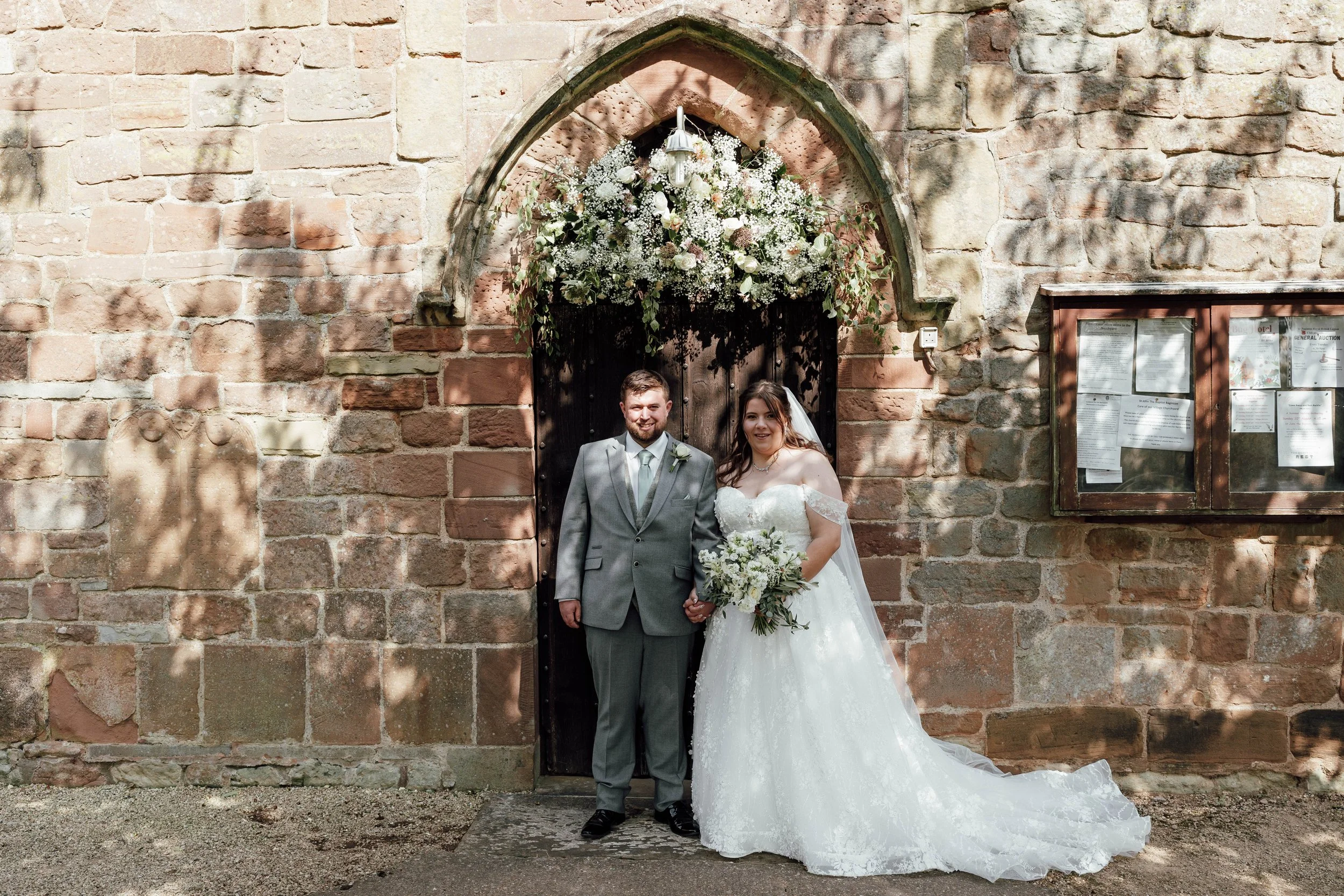 A bride and groom standing outside a church entrance decorated with flowers, holding hands and smiling.