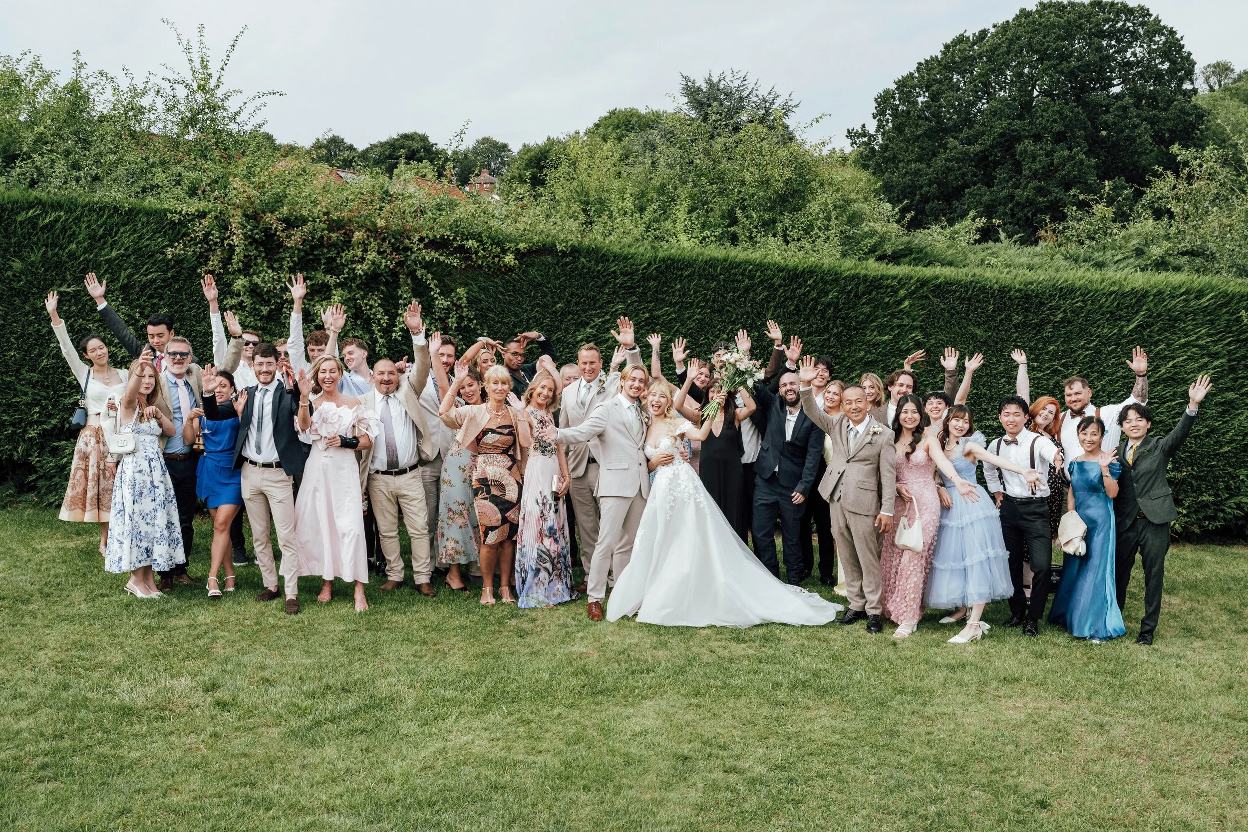A large group of wedding guests gathers outdoors on a grassy lawn, raising their hands in celebration around a bride and groom who are smiling. The bride is holding a bouquet of flowers, and the groom is in a light-colored suit. The group is dressed 