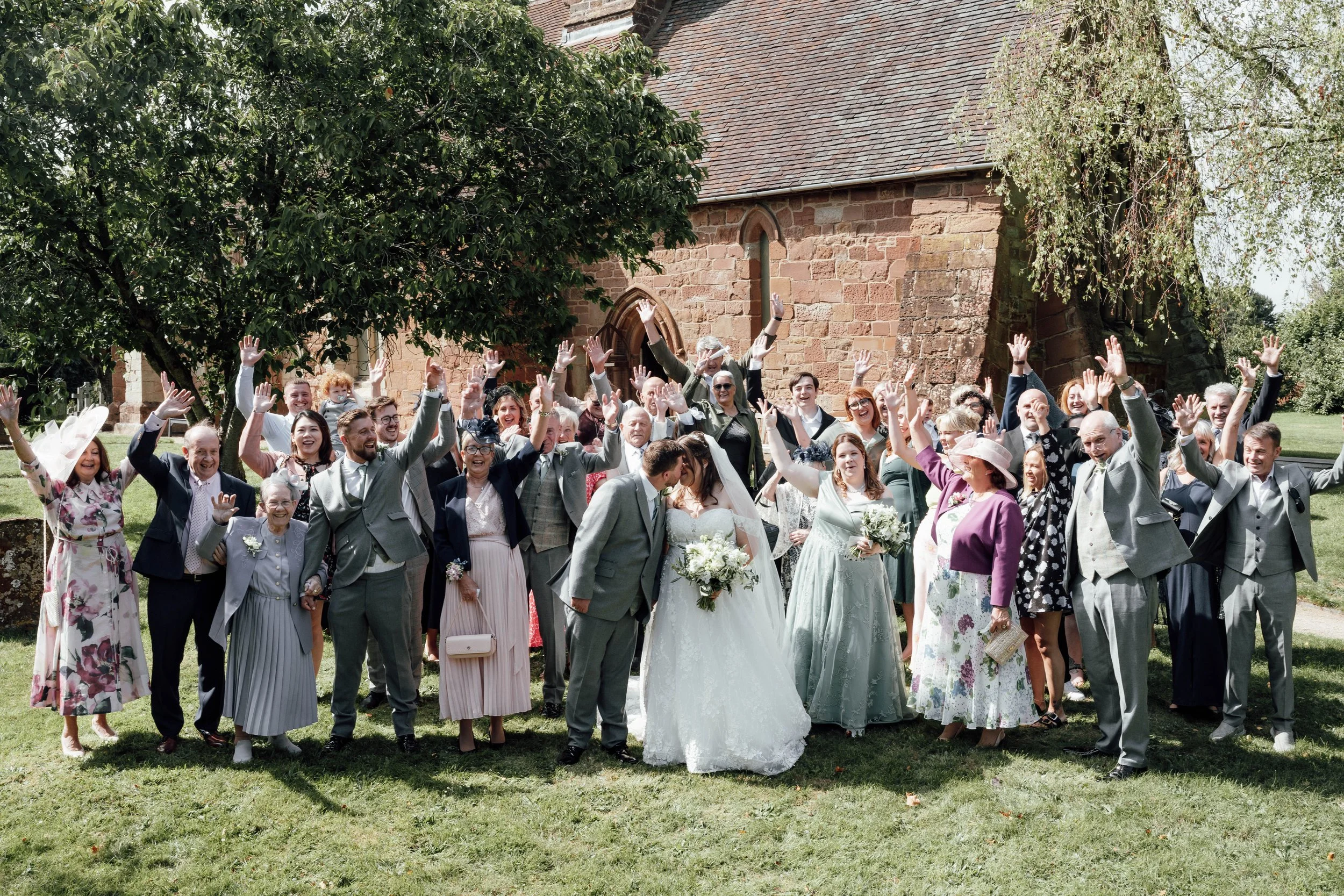 A large group of people celebrating outdoors near a brick church, with some raising their hands, and two people in the center, likely the bride and groom, kissing while surrounded by friends and family.