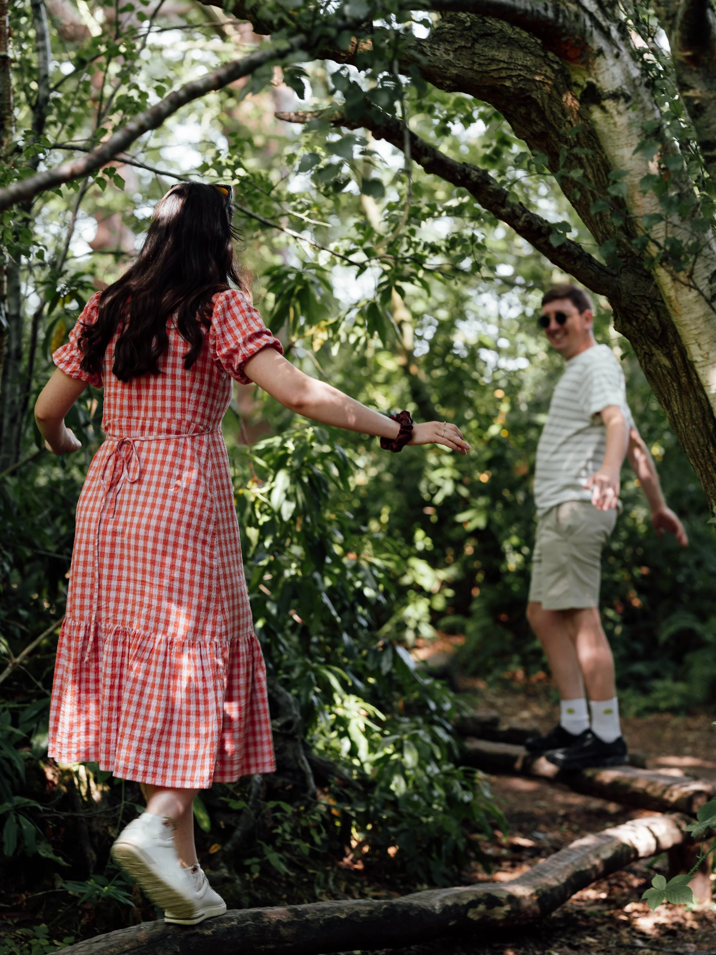 A woman in a red checkered dress walking on a log in the woods while a man in shorts and a striped shirt balances on a log nearby.