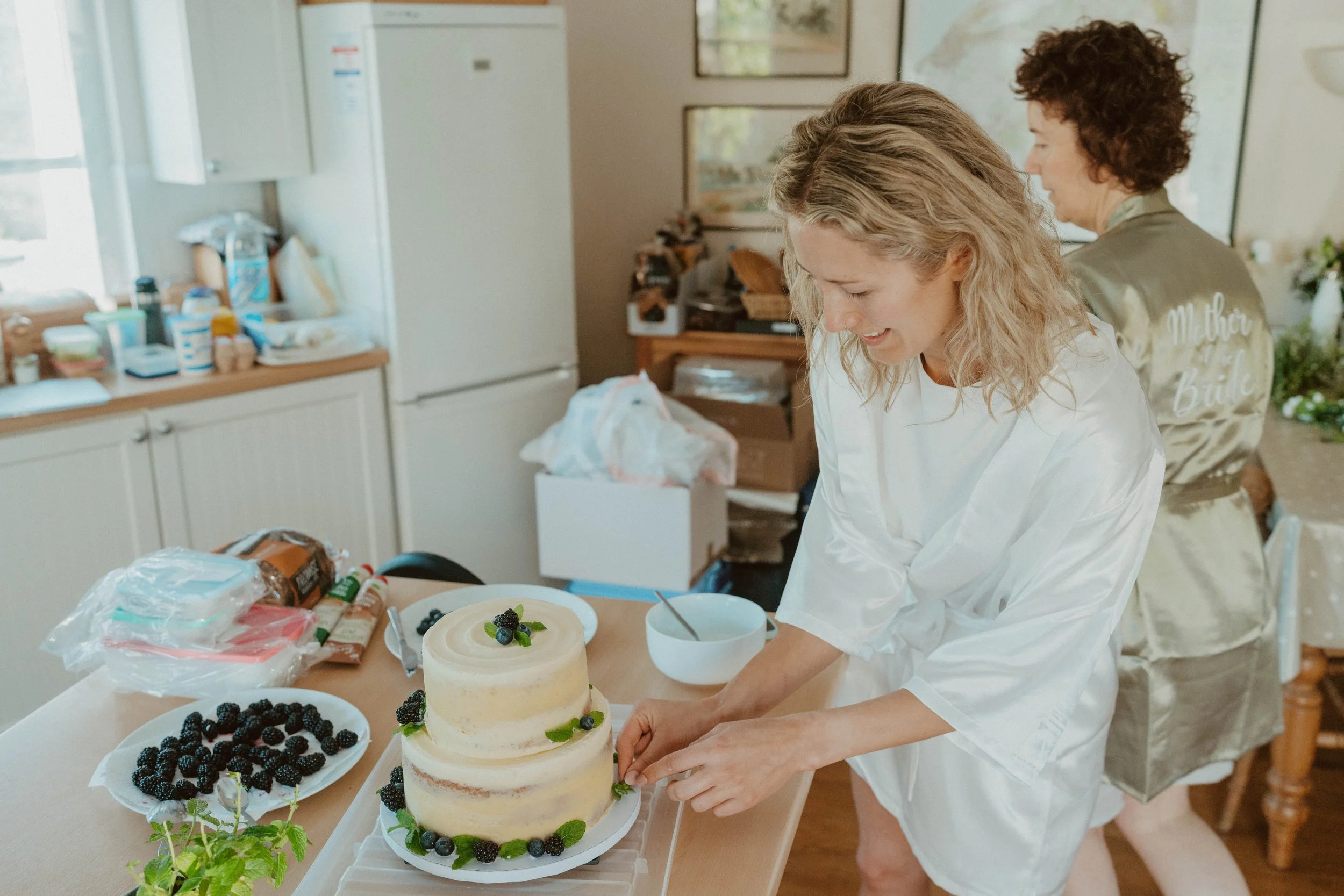 A woman in a white robe is decorating a two-tiered cake with blackberries and mint leaves, with other berries and baking supplies on the table.