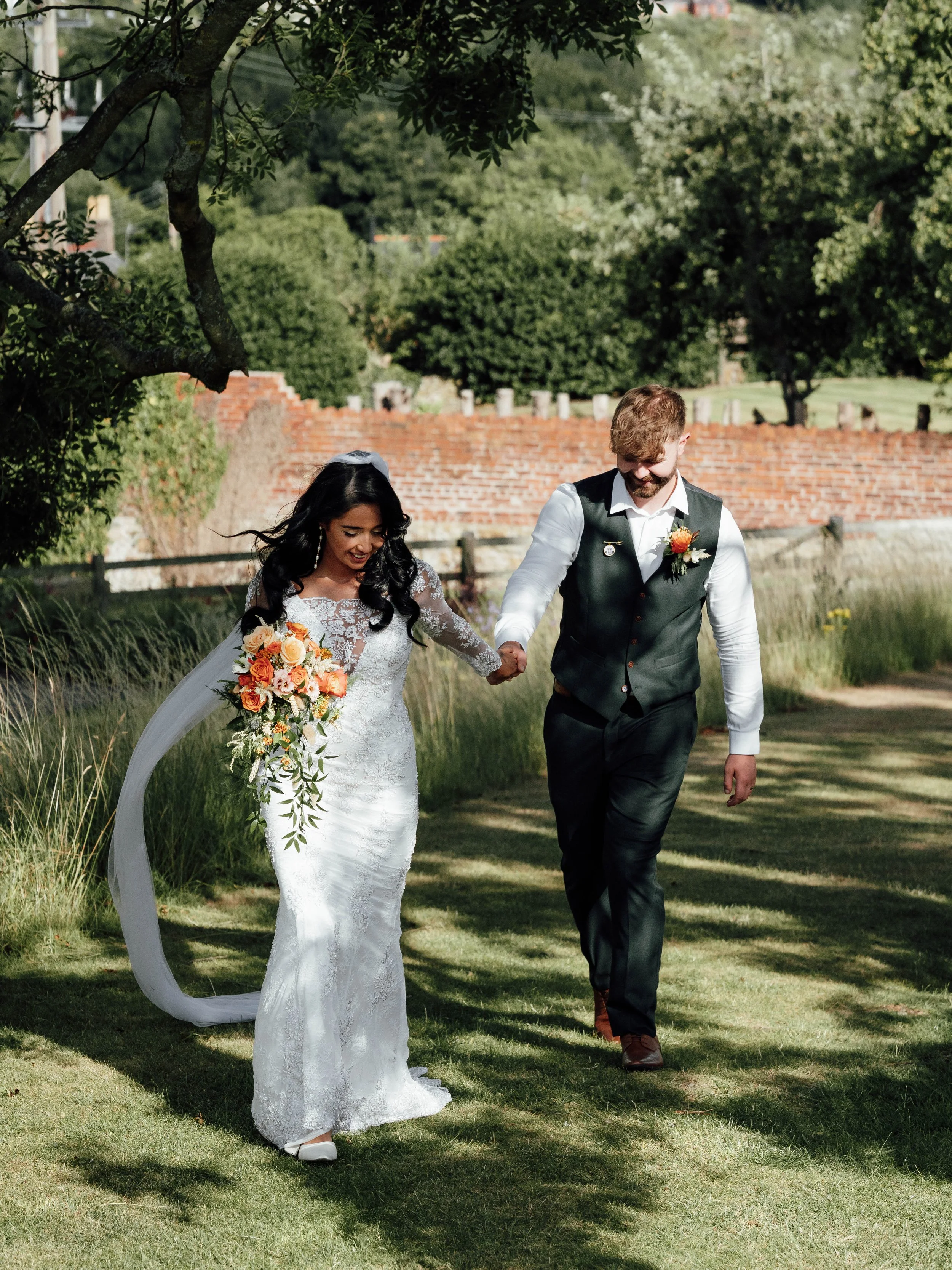 A bride and groom walking hand-in-hand outdoors on their wedding day, with the bride wearing a white lace wedding gown and holding a bouquet of orange and pink flowers, and the groom dressed in a dark vest and white shirt.