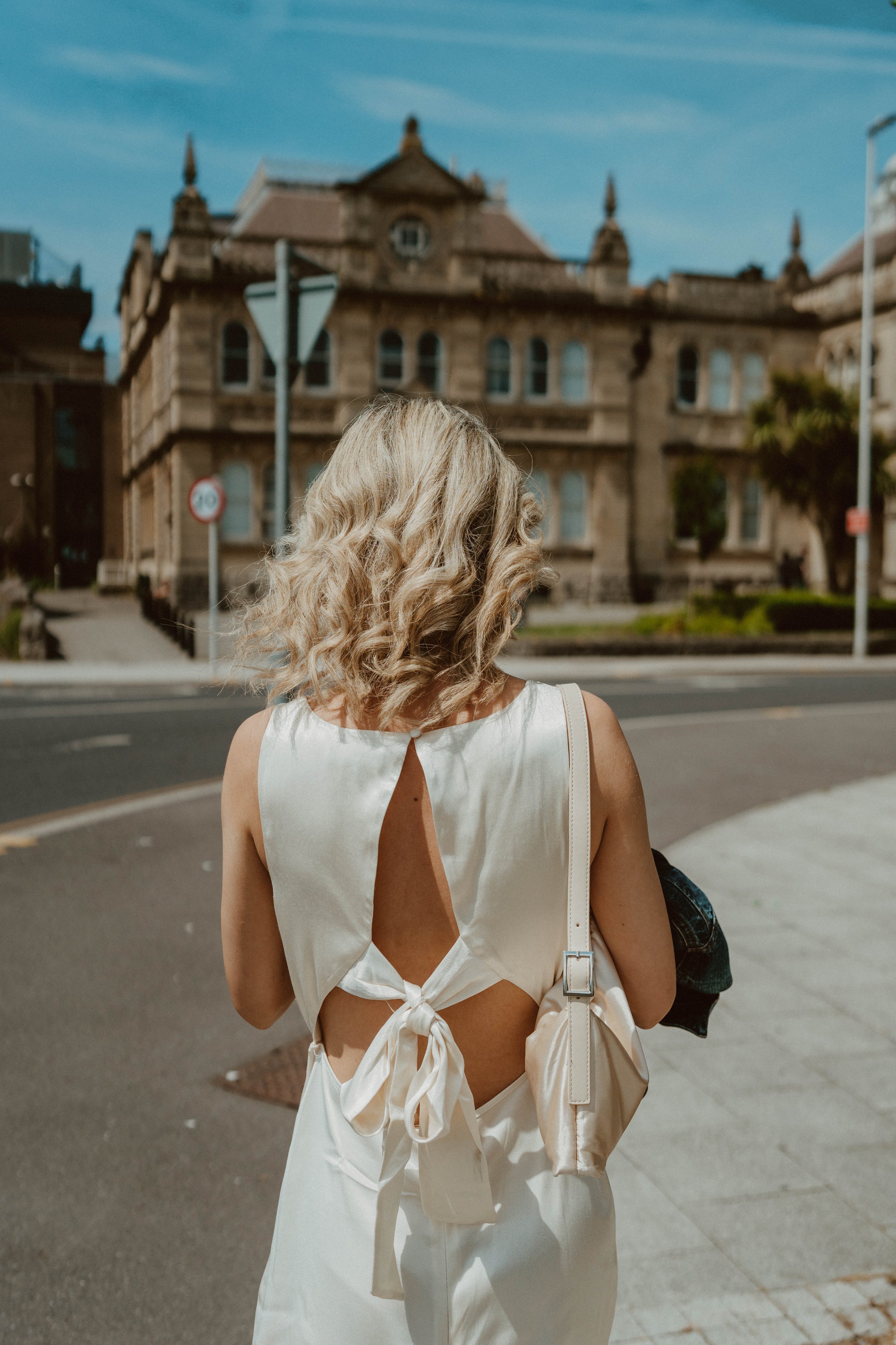 A woman with blonde curly hair stands on a city sidewalk facing away, wearing a white sleeveless dress with an open back tied with a bow, carrying a light-colored handbag and holding a black jacket.