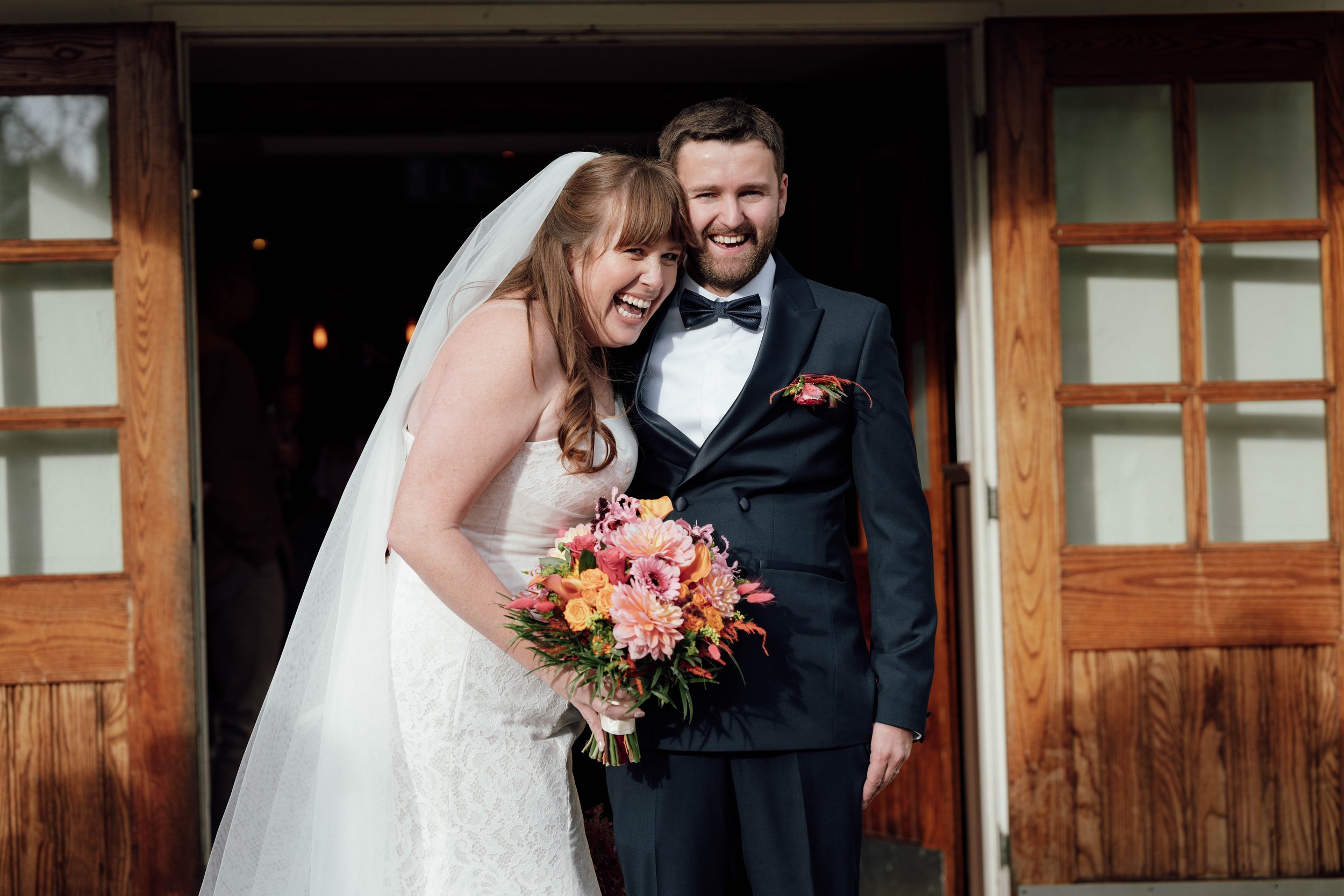 A bride and groom smiling and posing for a photo outside a wooden building, with the bride holding a colorful bouquet of flowers.