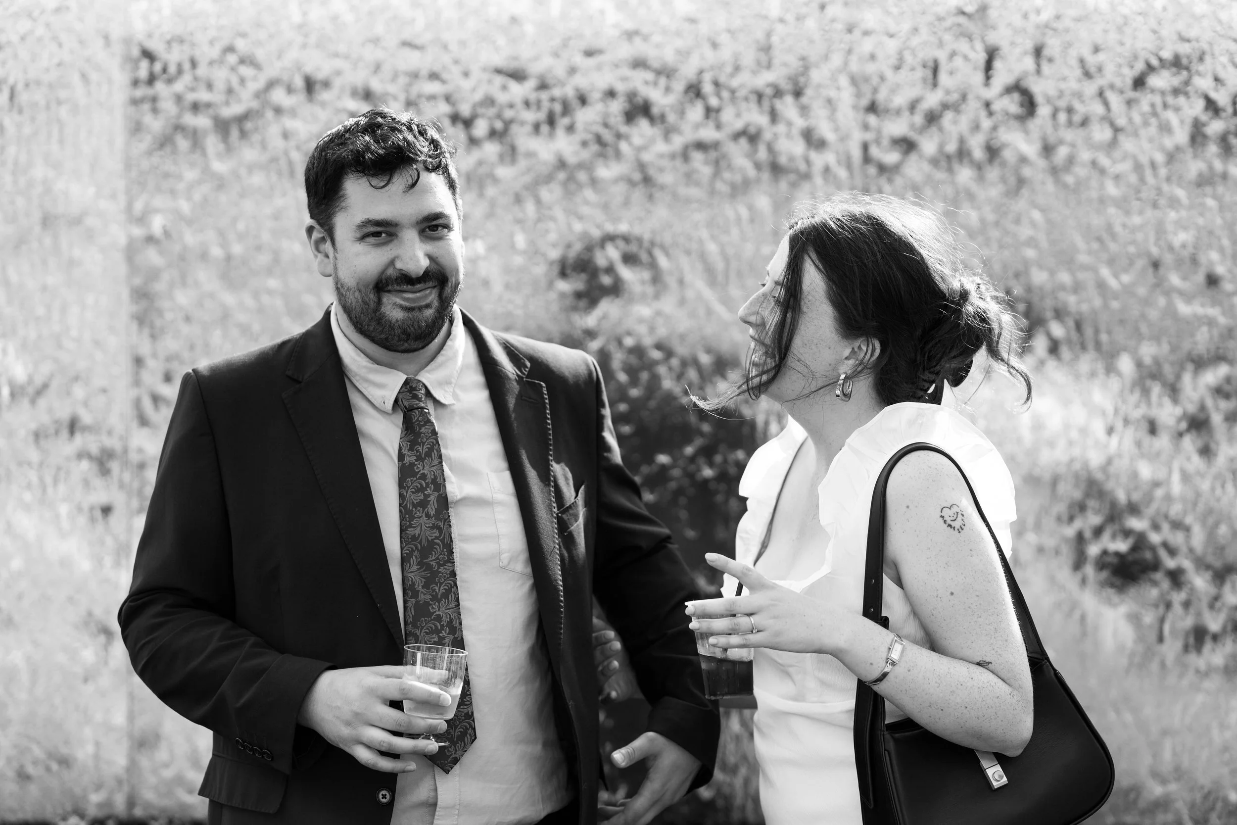 A black and white photo of a man in a suit and a woman in a white dress with freckles, standing outdoors with trees in the background. They are smiling and holding drinks, engaged in conversation.