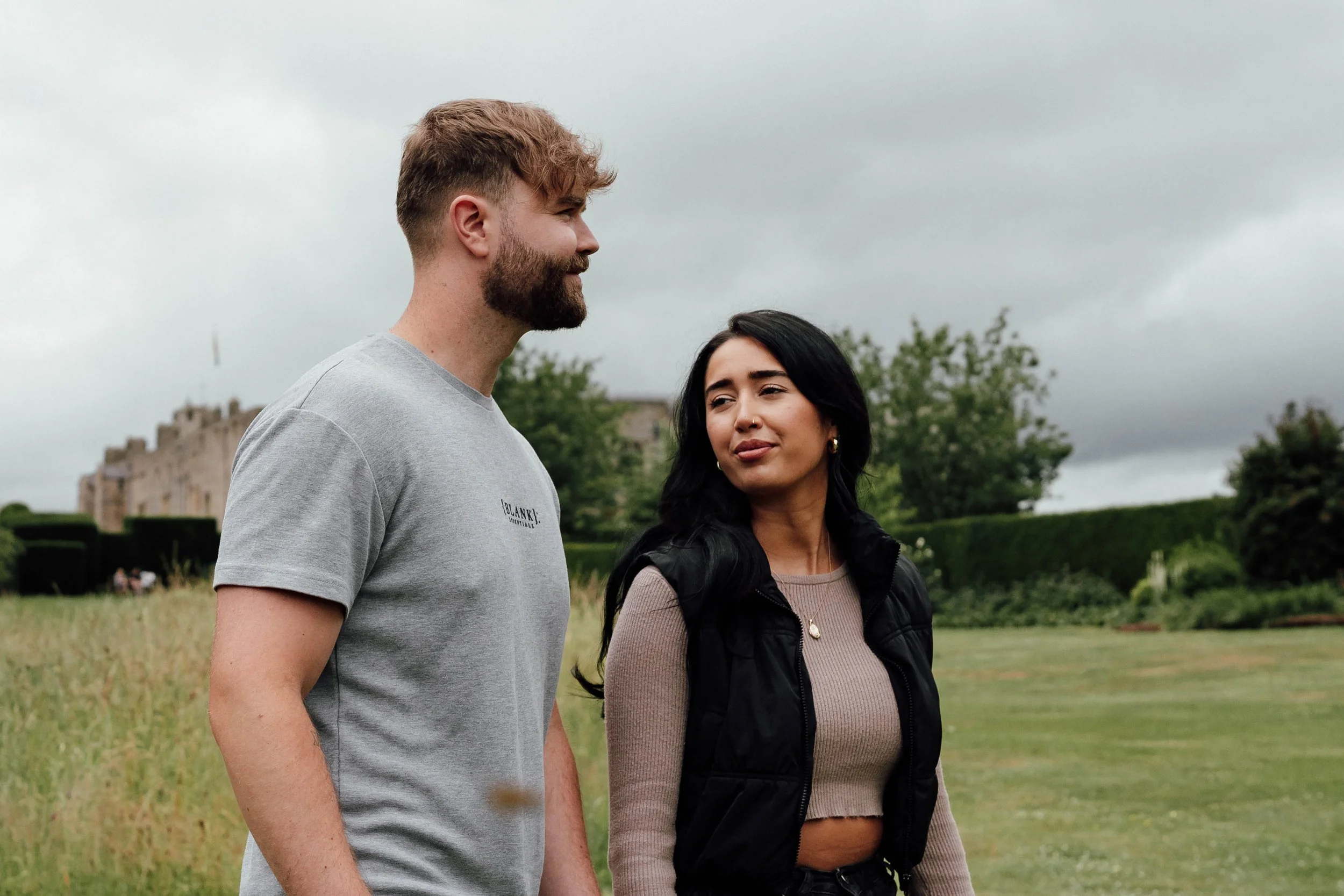 A young man and woman standing outdoors in a grassy area with trees and a cloudy sky behind them.