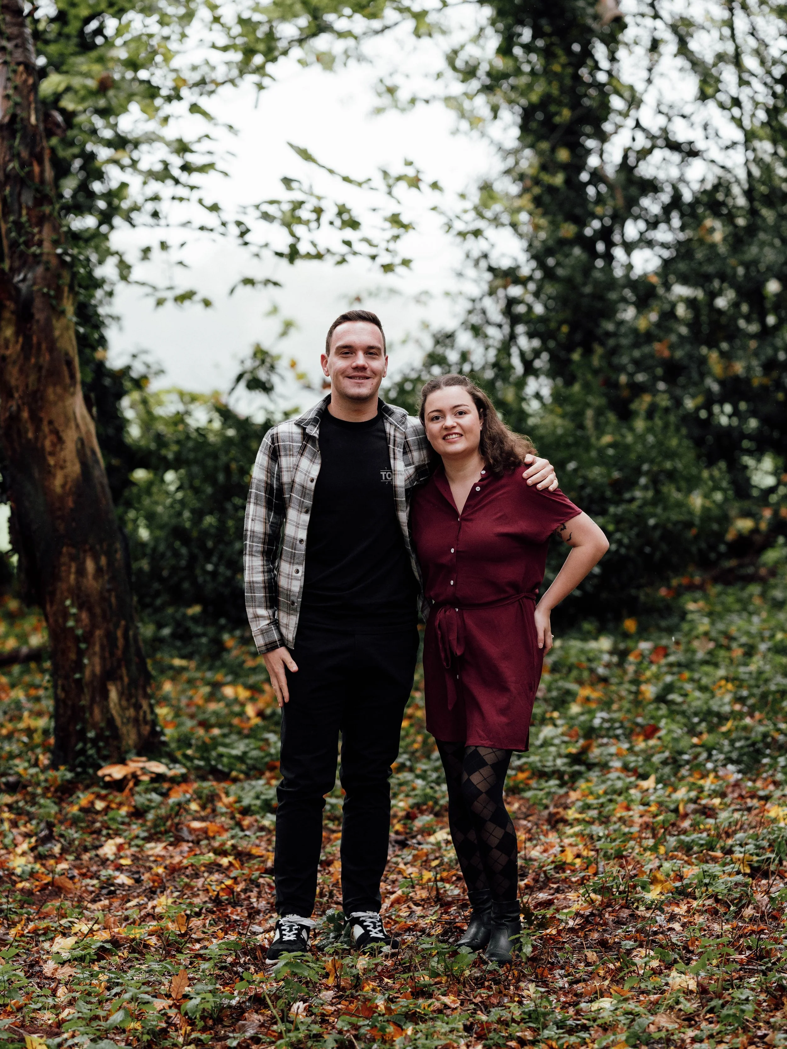 A young man and woman standing together outdoors in a wooded area, smiling at the camera, with the woman resting her hand on the man's shoulder.