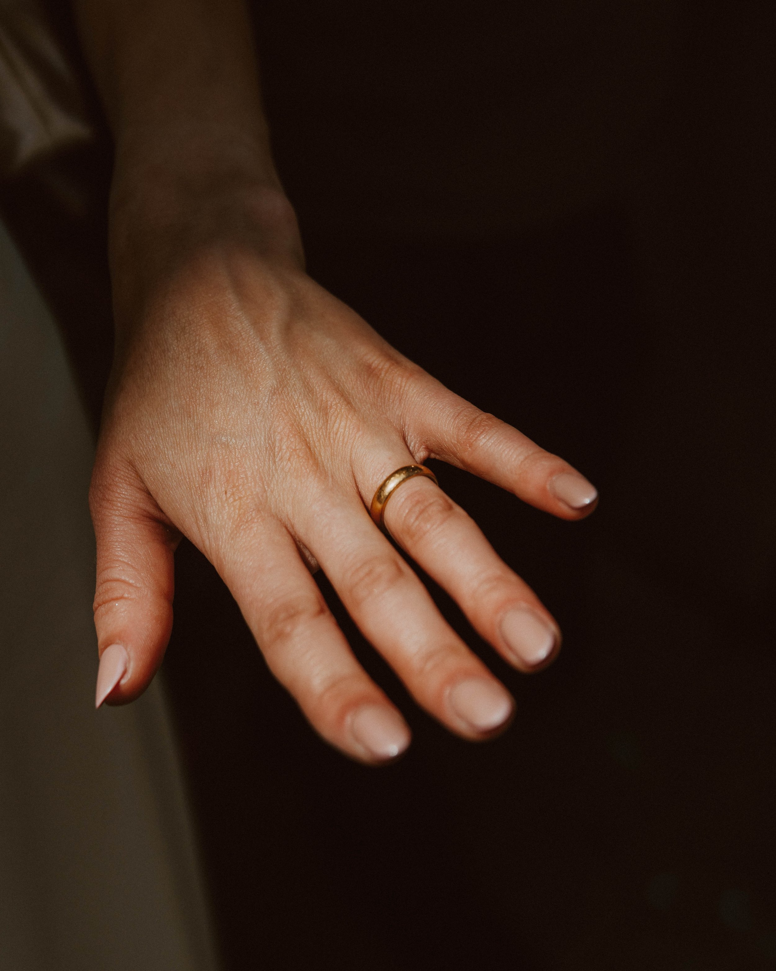 Close-up of a woman's left hand wearing a gold wedding band, palm down, against a dark background.