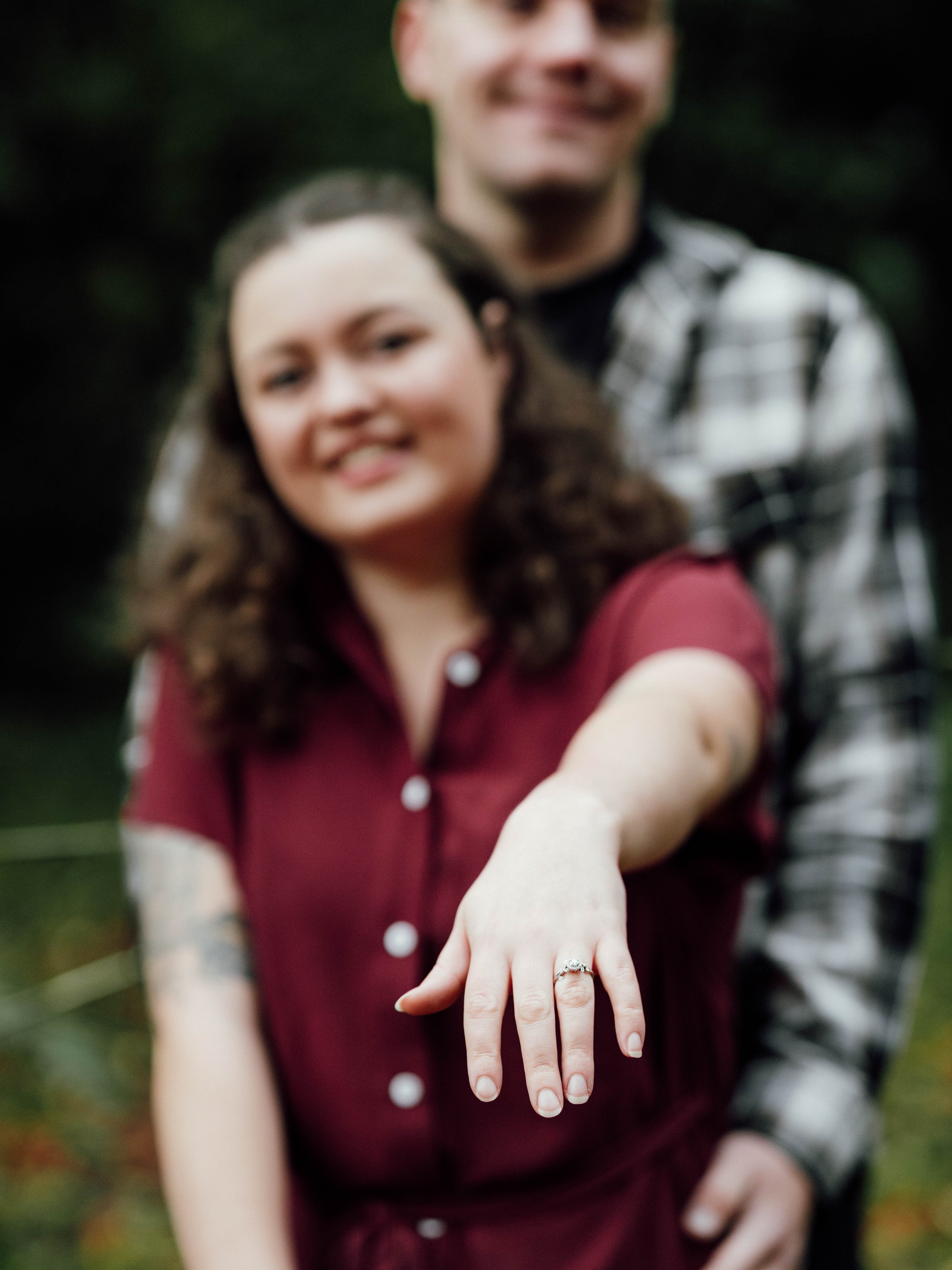 A woman shows off her engagement ring while smiling, with a man standing behind her, outdoors in a natural setting.