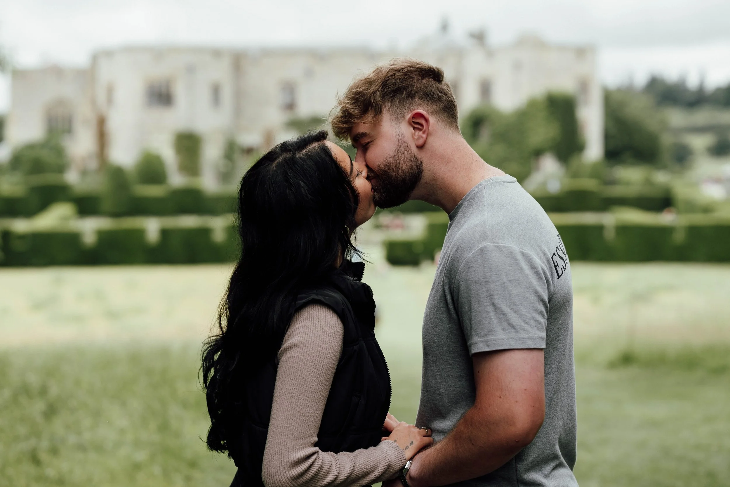A couple sharing a kiss outdoors in front of a grassy landscape and a large, historic building in the background.