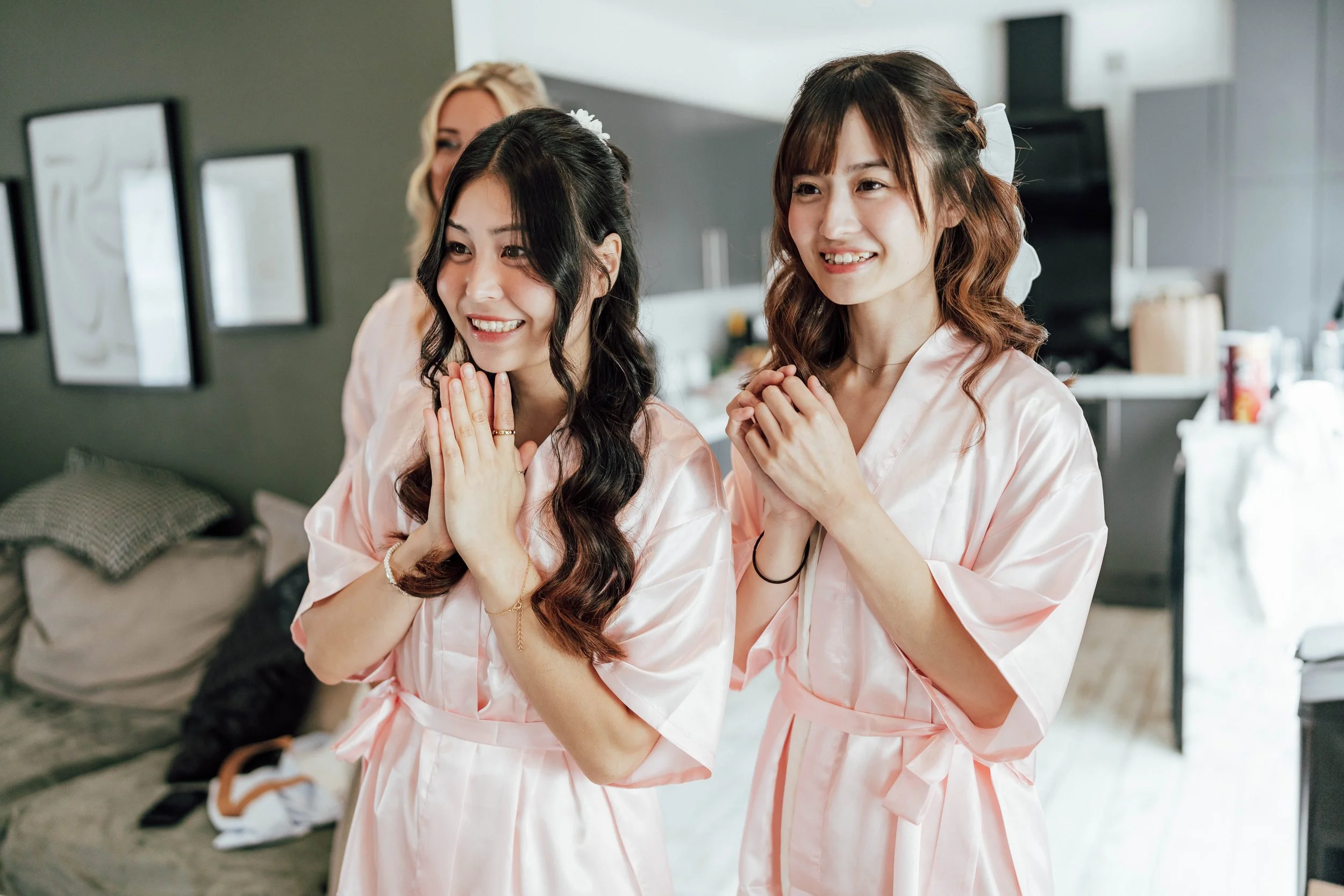 Two women in pink satin robes with hands clasped, smiling, and looking at something outside the frame, in a room with a couch, picture frames, and kitchen in the background.