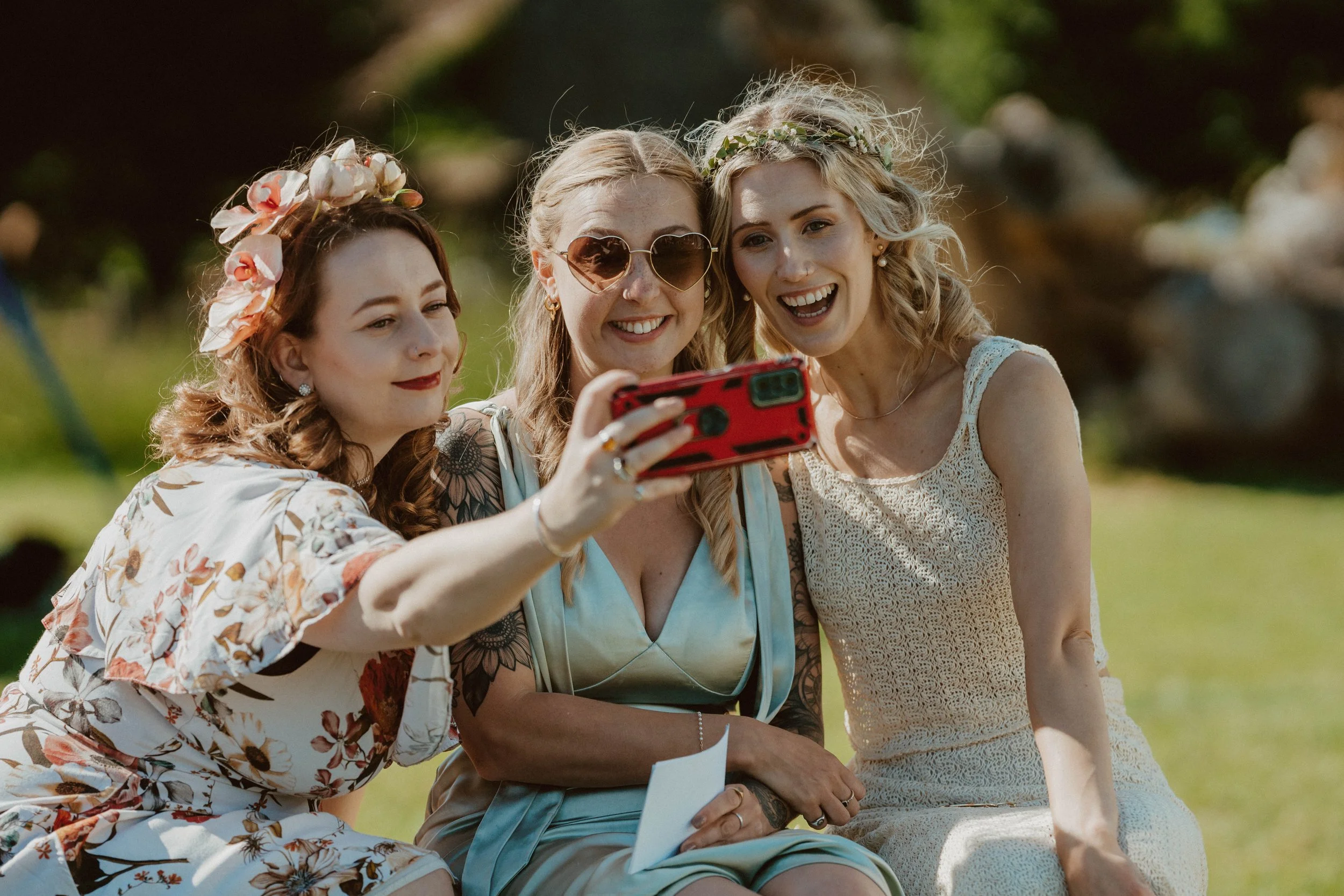 Three women taking a selfie outdoors at a wedding or celebration, with green trees and grass in the background.