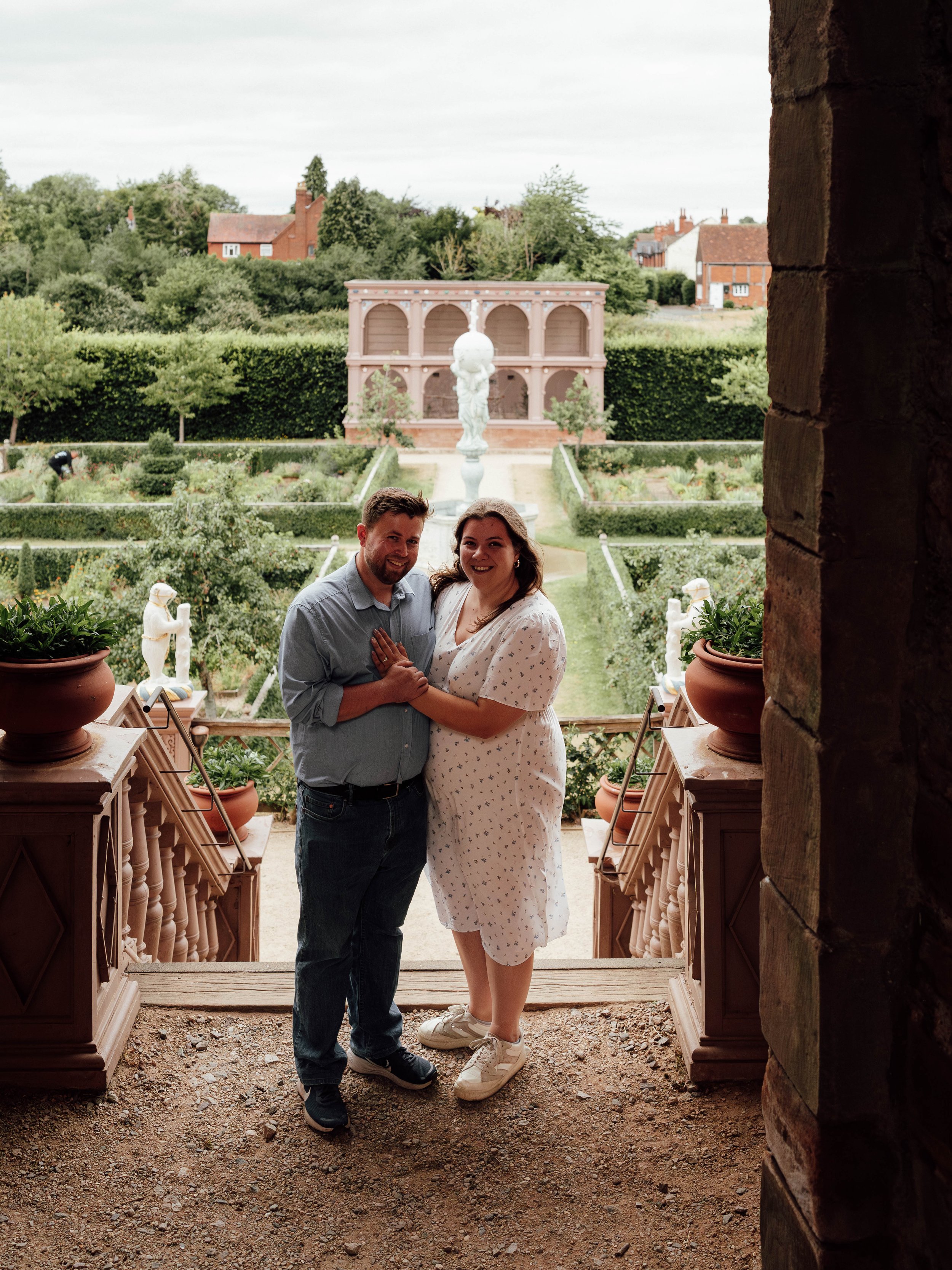 A couple standing on a porch with a garden and fountain in the background.