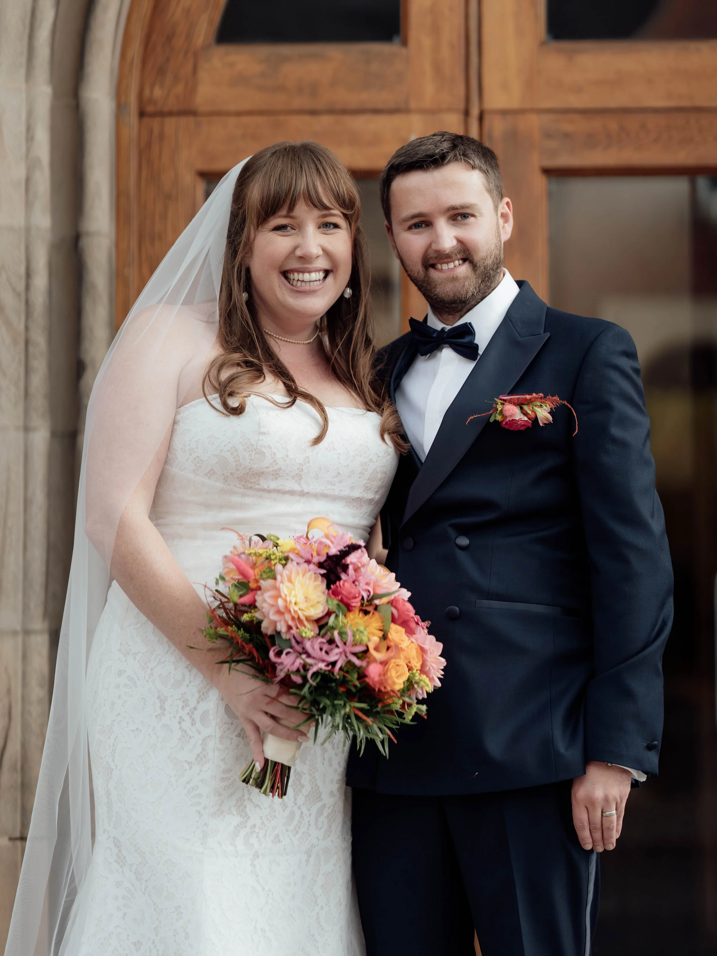 A bride in a white lace wedding dress holding a colorful bouquet of flowers and a groom in a black tuxedo with a bow tie, standing in front of a wooden door, smiling.