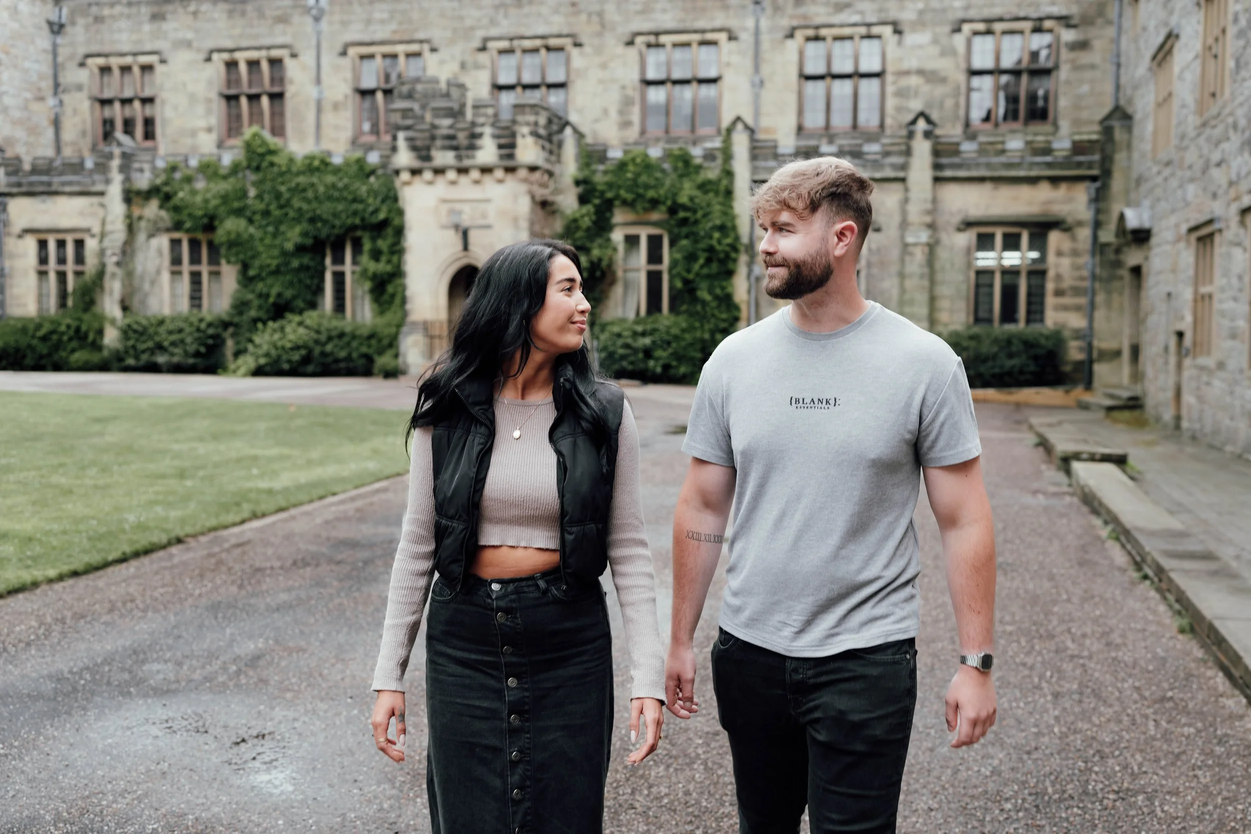 A young woman and man walk together on a campus or historic building background, looking at each other, holding hands, with grass, trees, and stone buildings visible behind them.