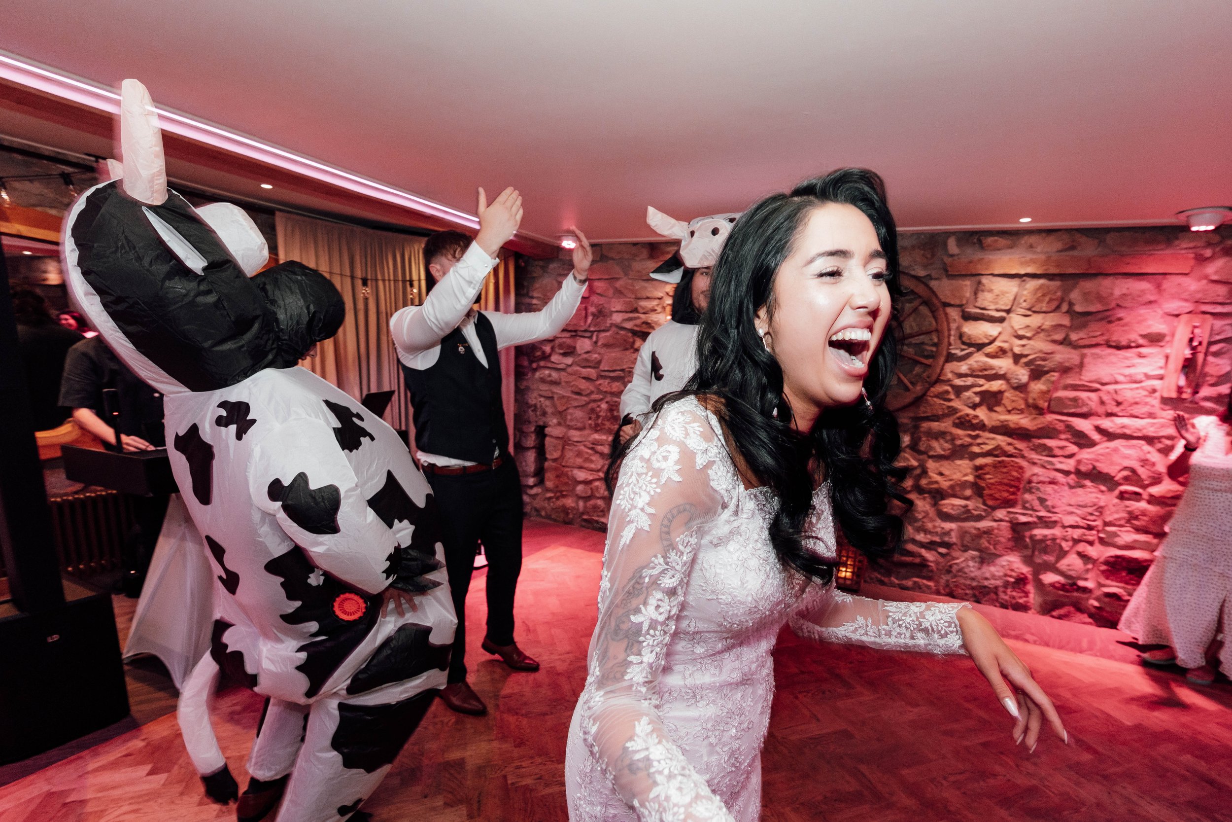 Woman in white lace dress laughing at a celebration with people in cow costumes in a decorated indoor venue.