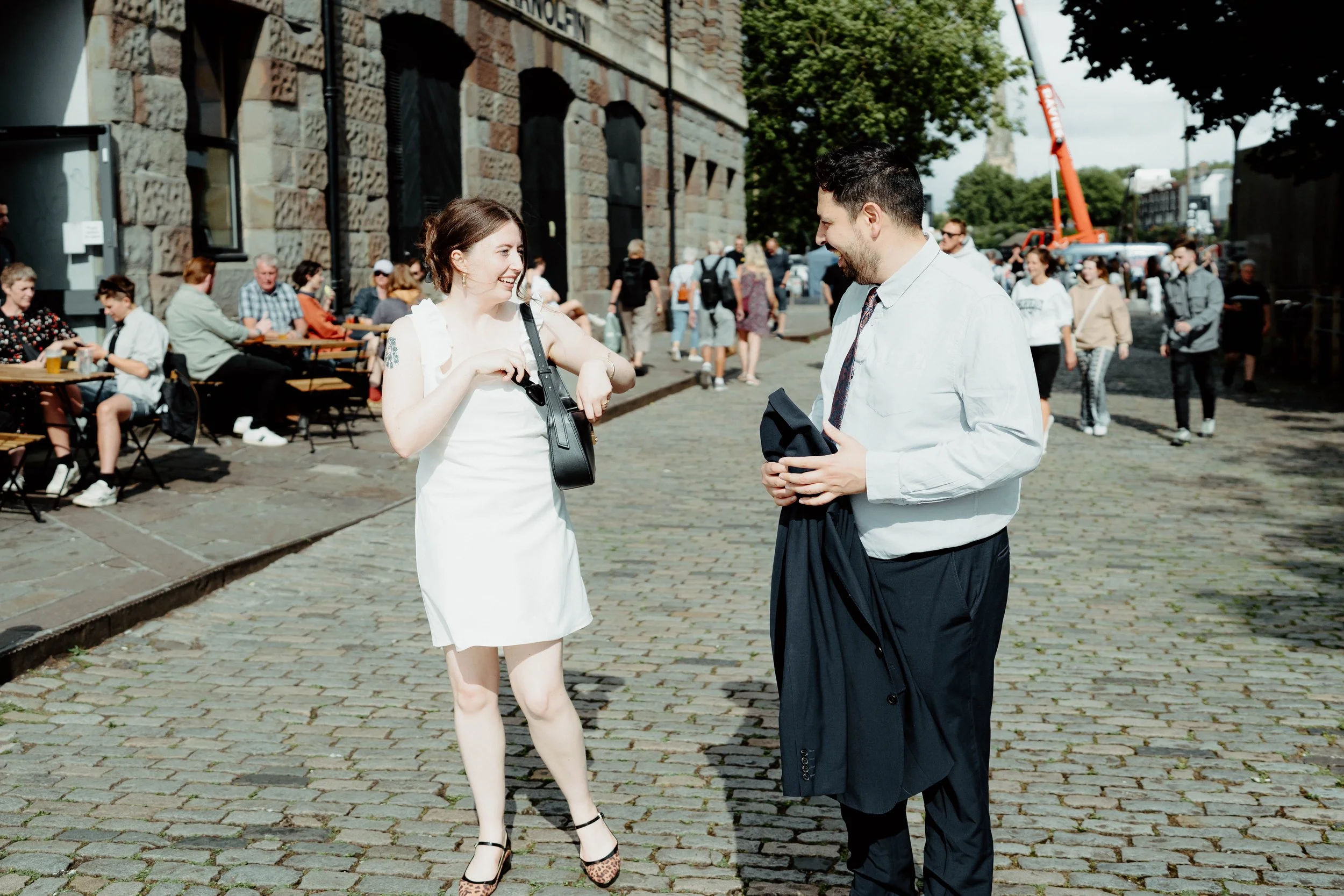 A young woman and man in formal attire smiling and talking on a cobblestone street outside a building, with people sitting at outdoor tables and walking in the background.