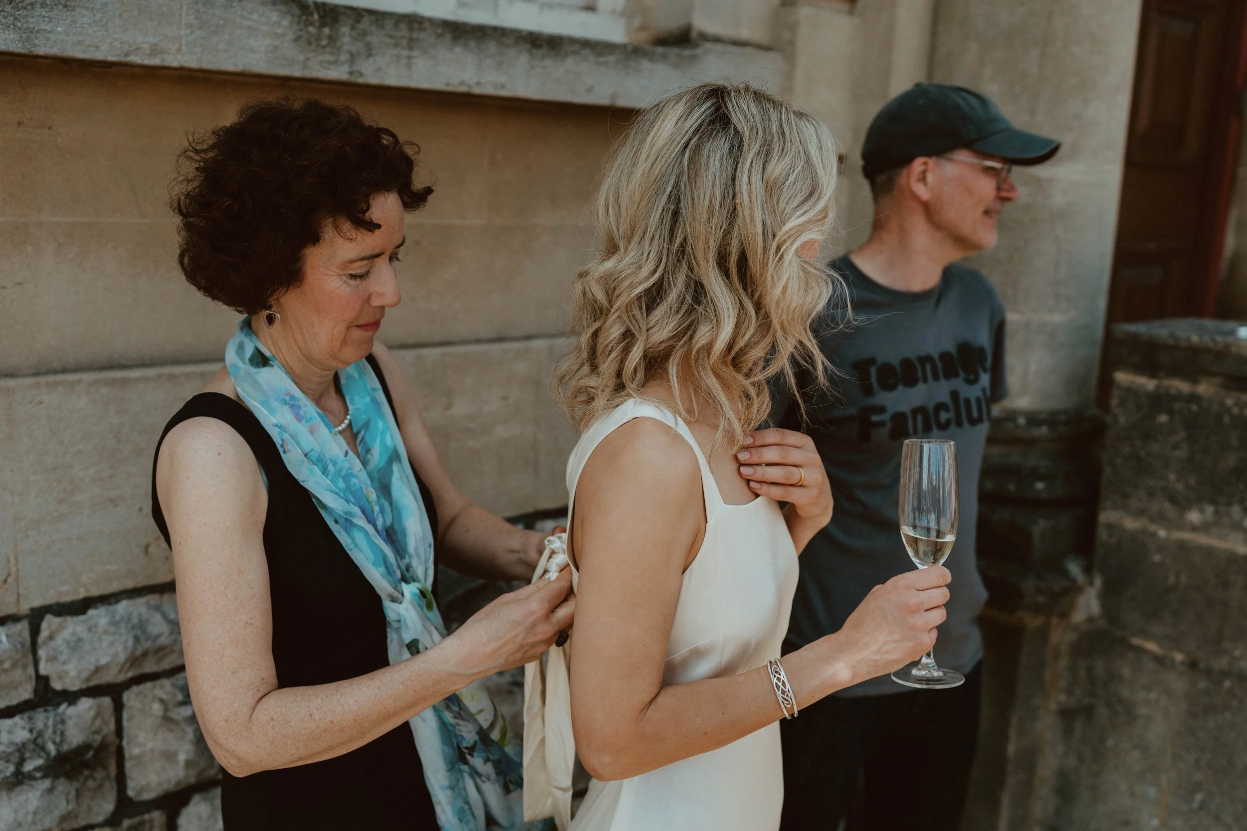 Three people standing outdoors against a stone wall. The woman on the left is fastening a bracelet on the woman in the middle, who is holding a glass of champagne. The man on the right is wearing a cap and dark T-shirt with the words 'Teenage Fanclub