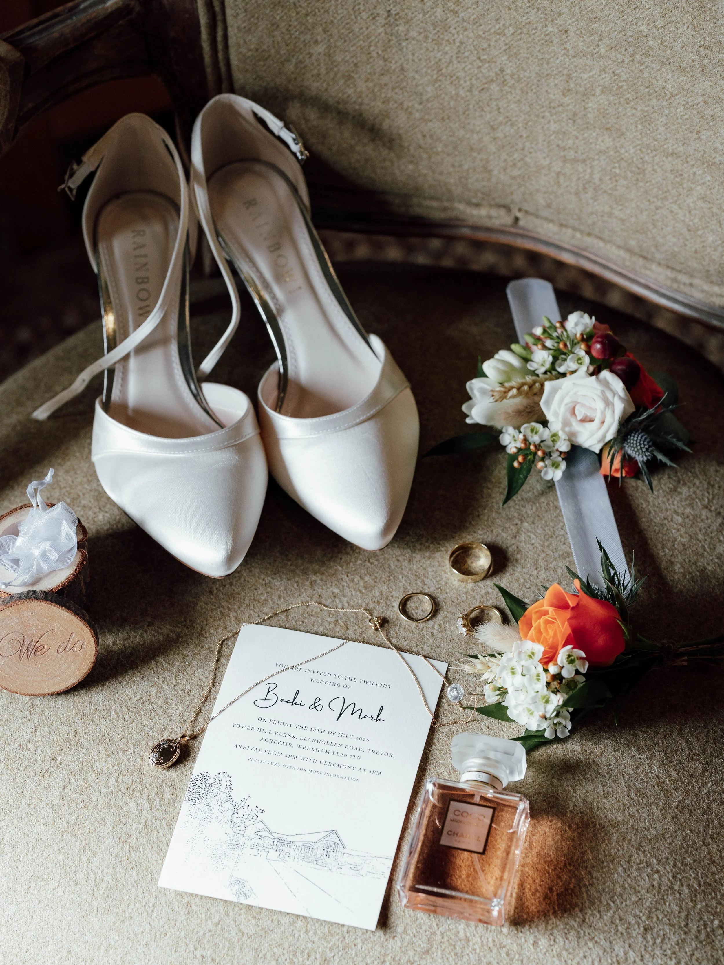 Wedding shoes, bouquet, wedding rings, perfume, and invitation on a beige fabric surface.