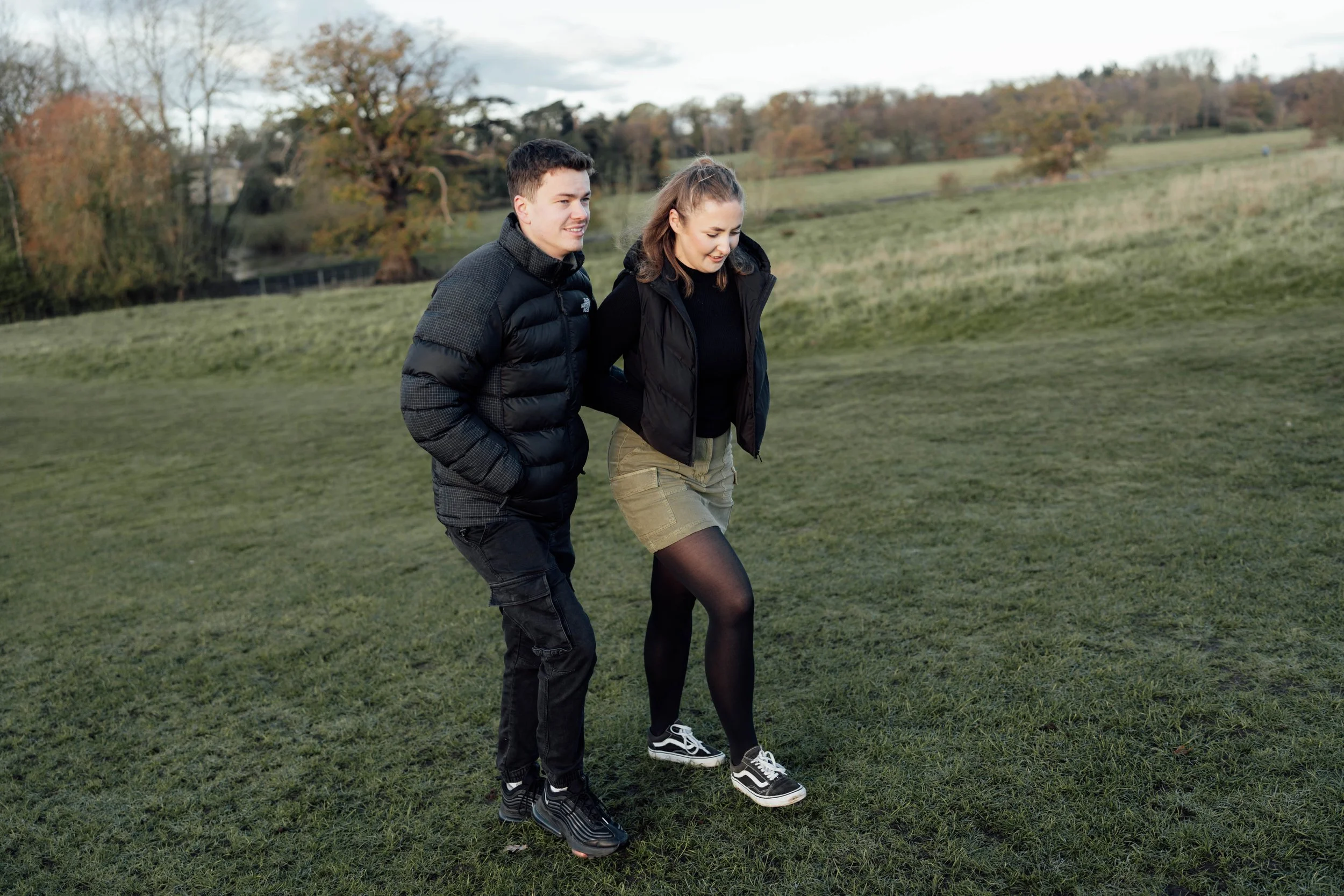 A young man and woman walking together on a grassy field in a park during fall, both dressed in black jackets and sneakers, with trees and hills in the background.