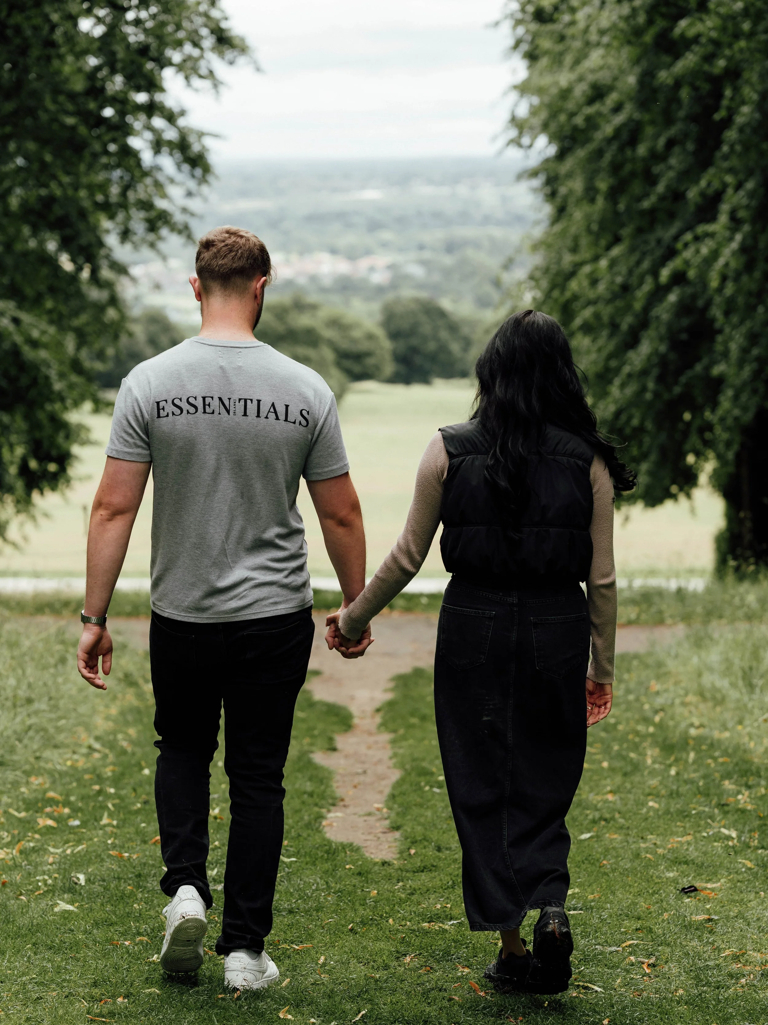 A couple holding hands and walking along a grassy path in a park with trees on both sides, overlooking a distant landscape.