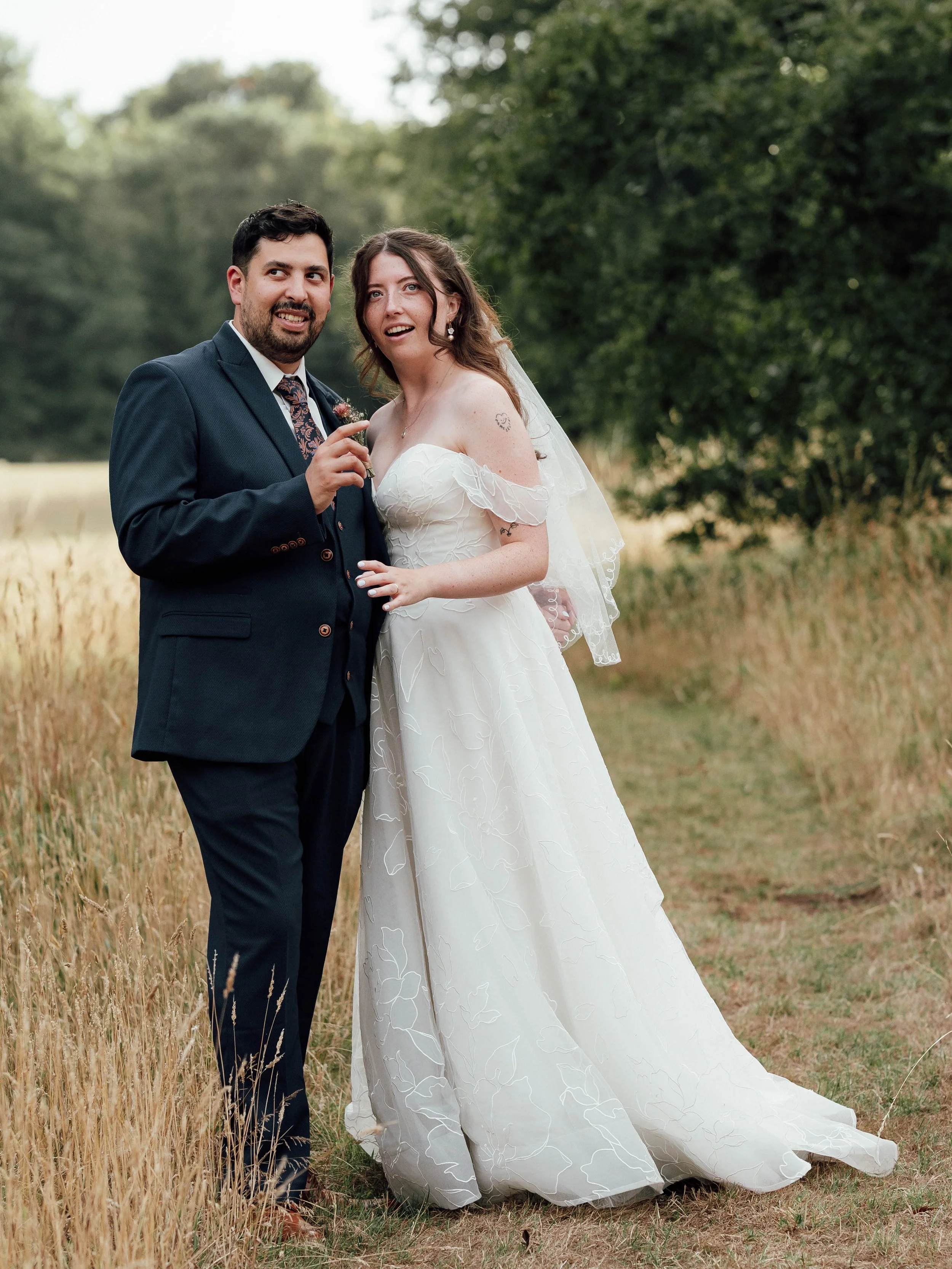 A bride and groom standing on a grassy path in a field, surrounded by trees, during their wedding photoshoot.
